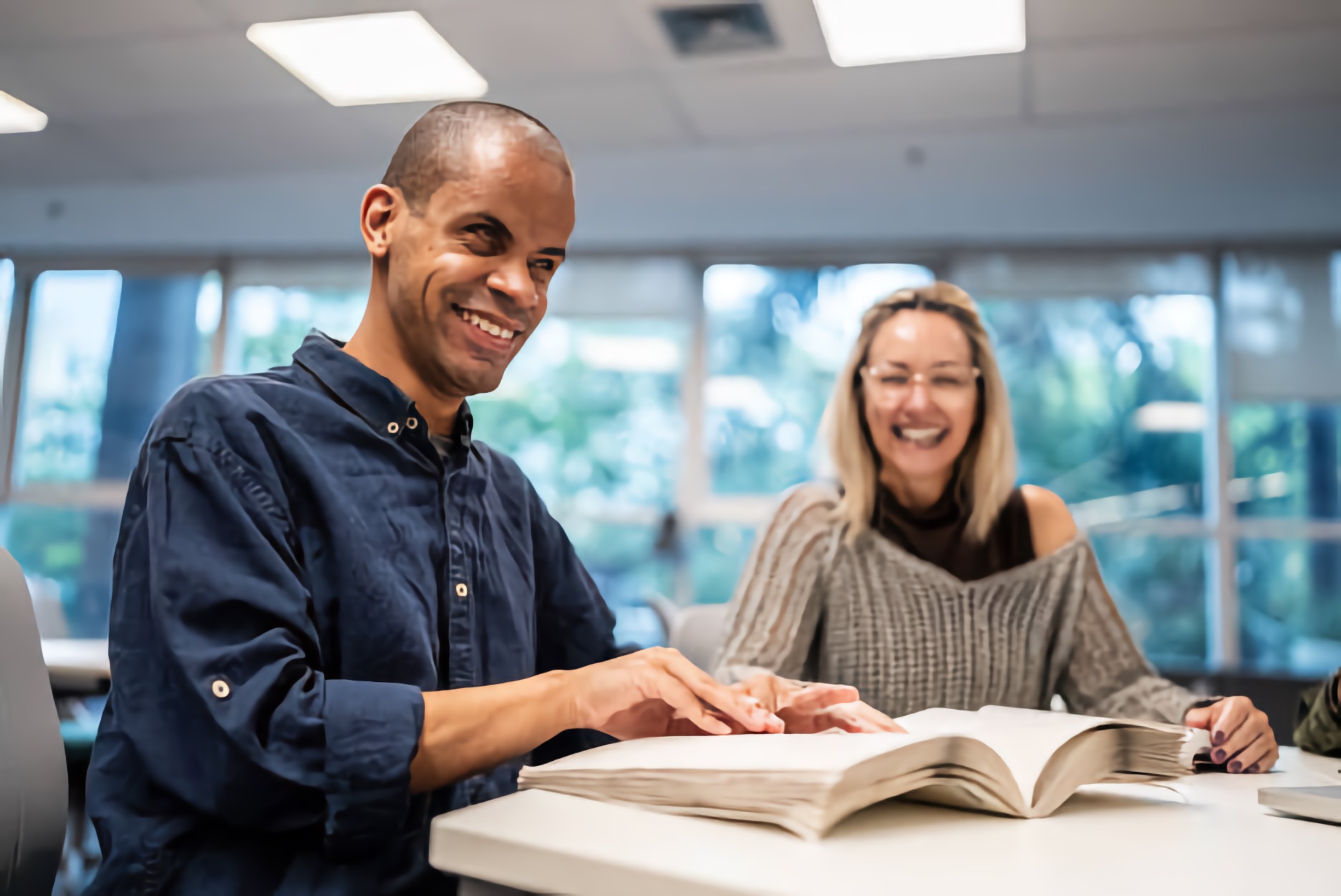 Blind Black gentleman reading a Braille book, with a smiling White woman beside him, at a gathering
