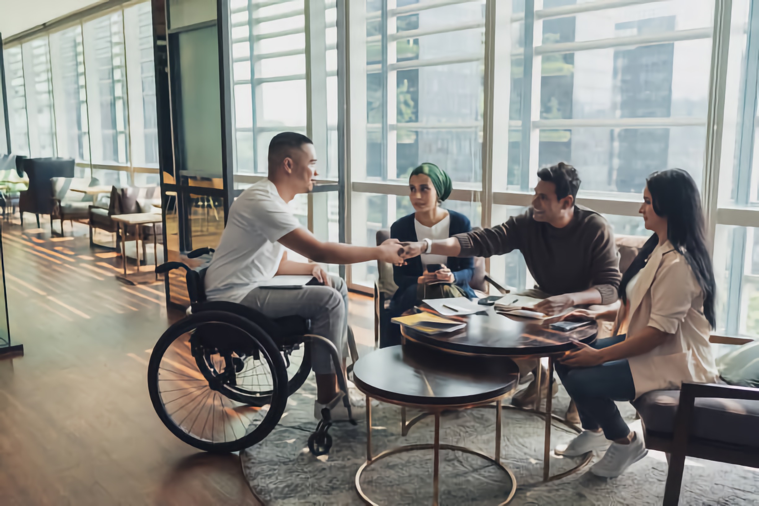Four individuals collaborate around a small round table in a public area, with a man using a wheelchair shaking hands with the man across from him