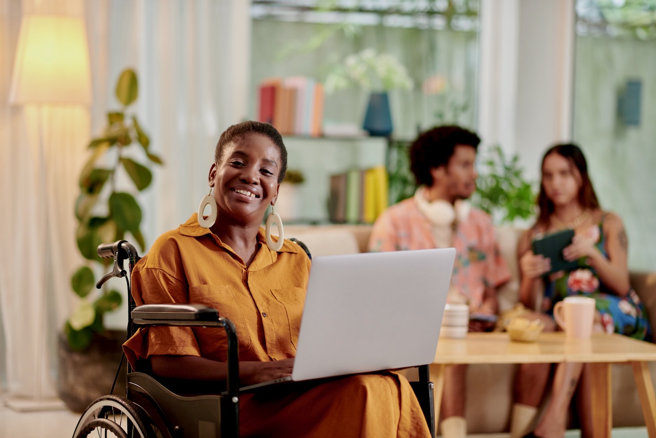A Black Woman who uses a wheelchair is smiling while using a Laptop Computer in a group meeting setting.