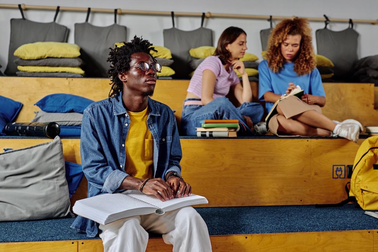 A blind black man sits on some risers with a study group, all engaged in reading activities, while he reads from a Braille book