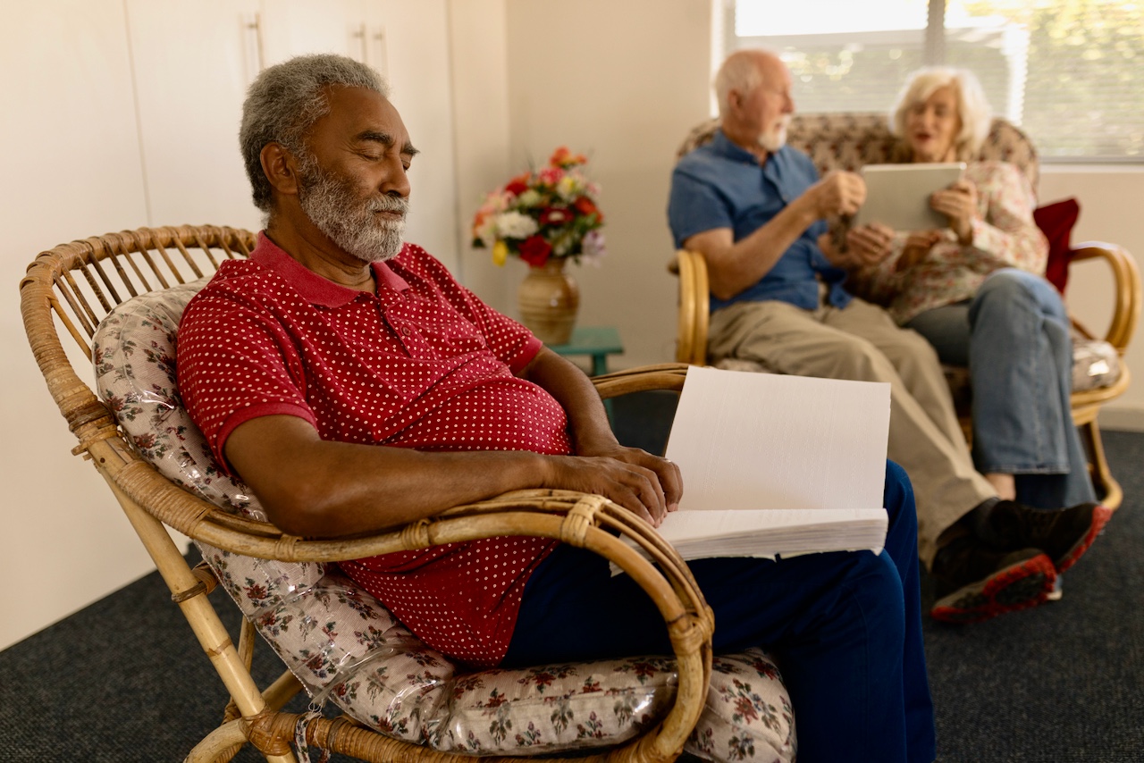 A retired blind black man sitting in a chair during a group meeting, reading from a Braille book with a serene look on his face, while an older couple follows along on an electronic tablet.