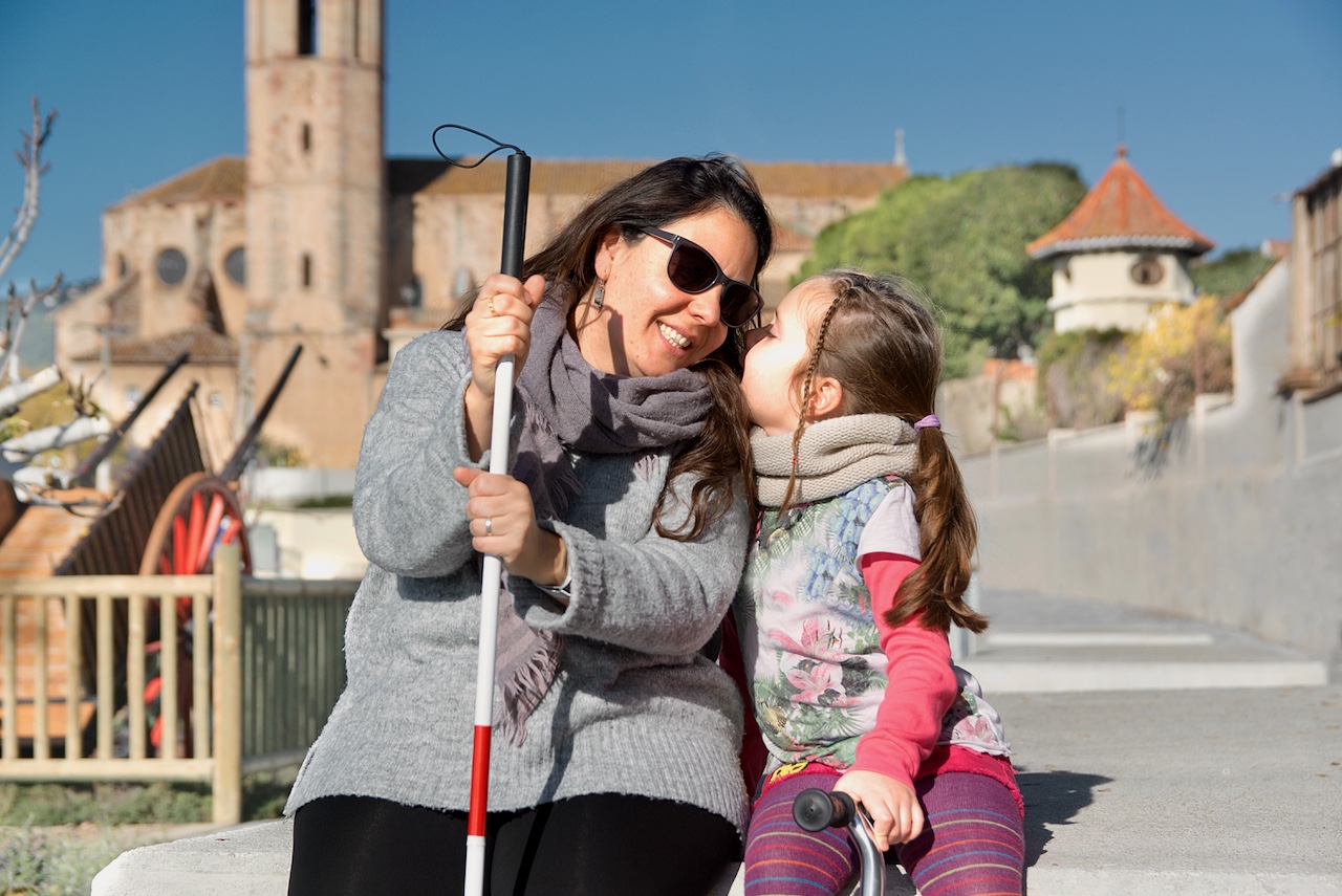 Blind mother with a white cane and her daughter enjoying a conversation while seated on a park bench, sharing smiles.