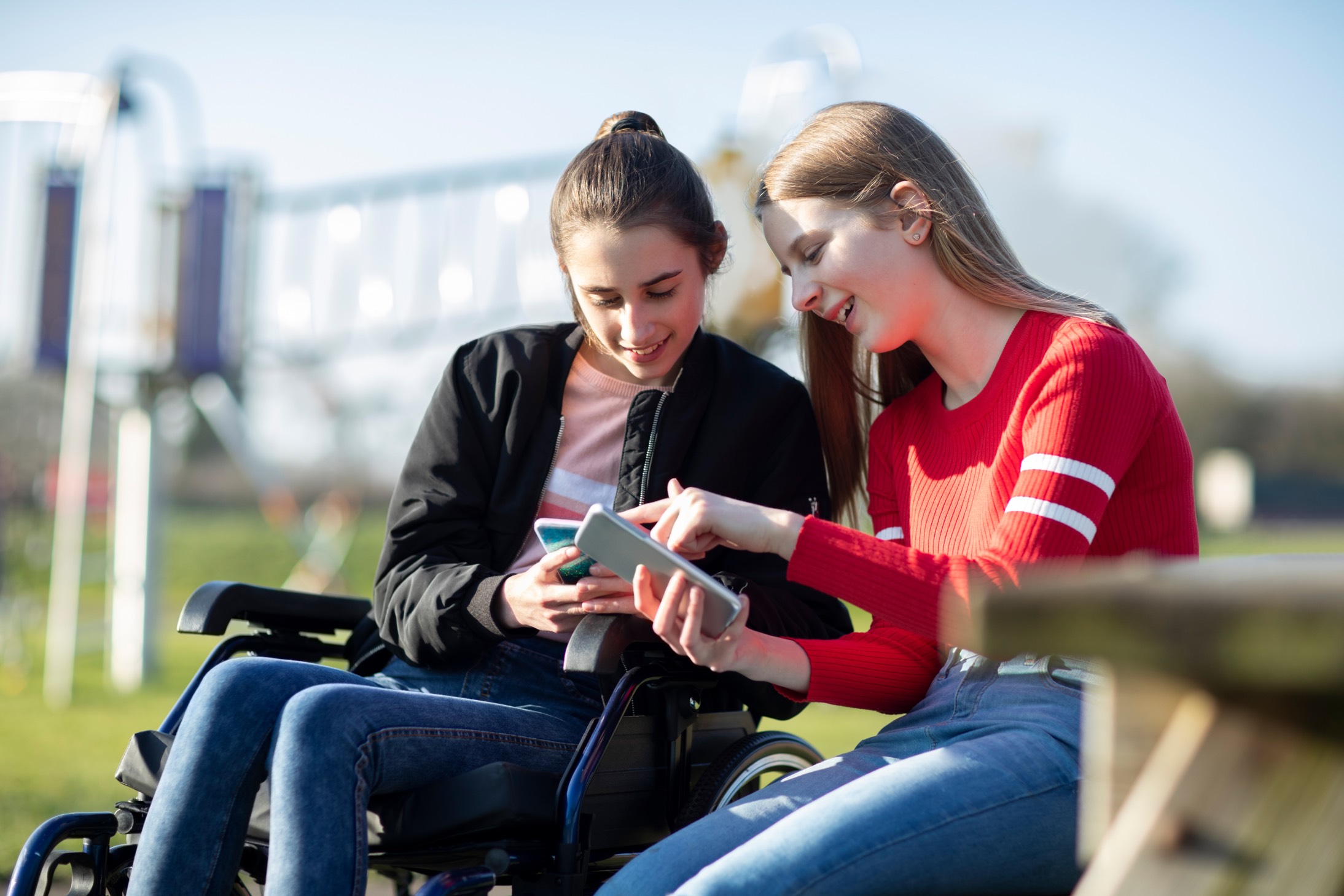 A teenage girl using a wheelchair comparing content on mobile phones with her friend in the park