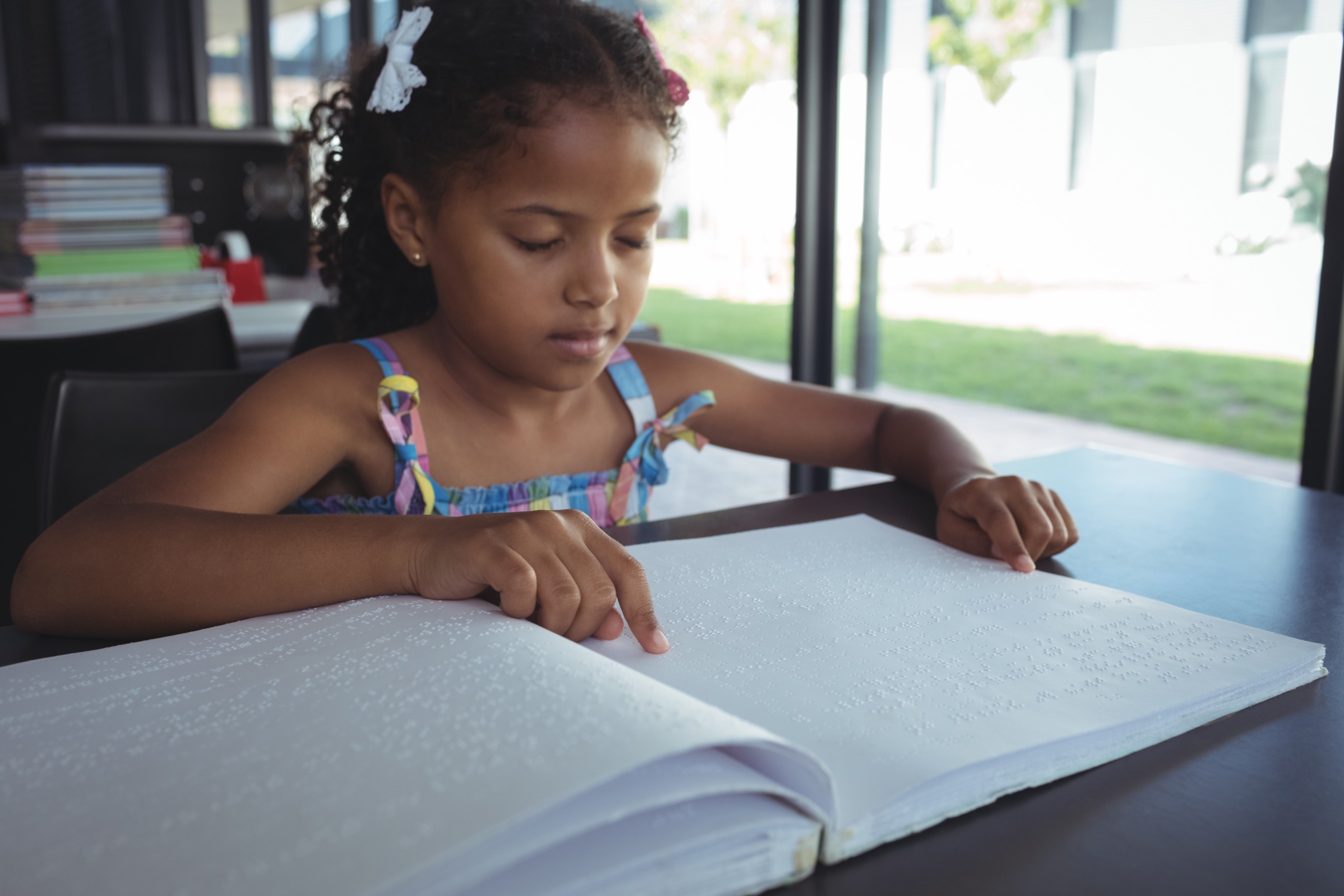 A young Black girl sits in a sunny room, immersed in reading Braille