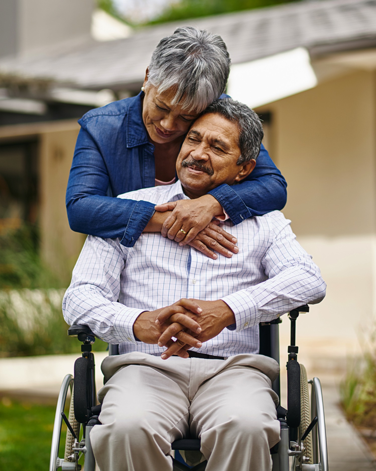 An older Hispanic couple smiles outdoors as the woman hugs the man, who uses a wheelchair, from behind around the shoulders