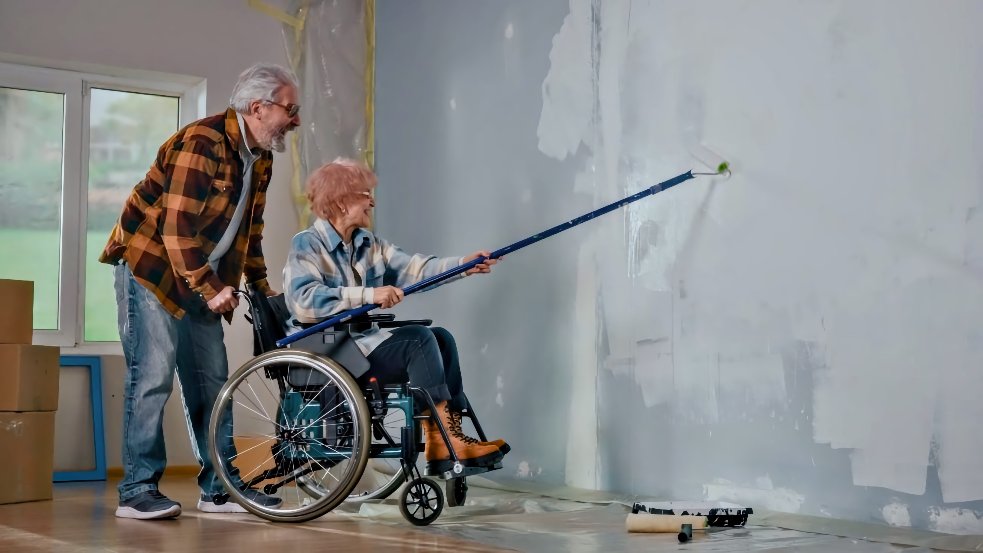 An older woman in a wheelchair using a paint roller on a wall, assisted by an elderly man adjusting her wheelchair.
