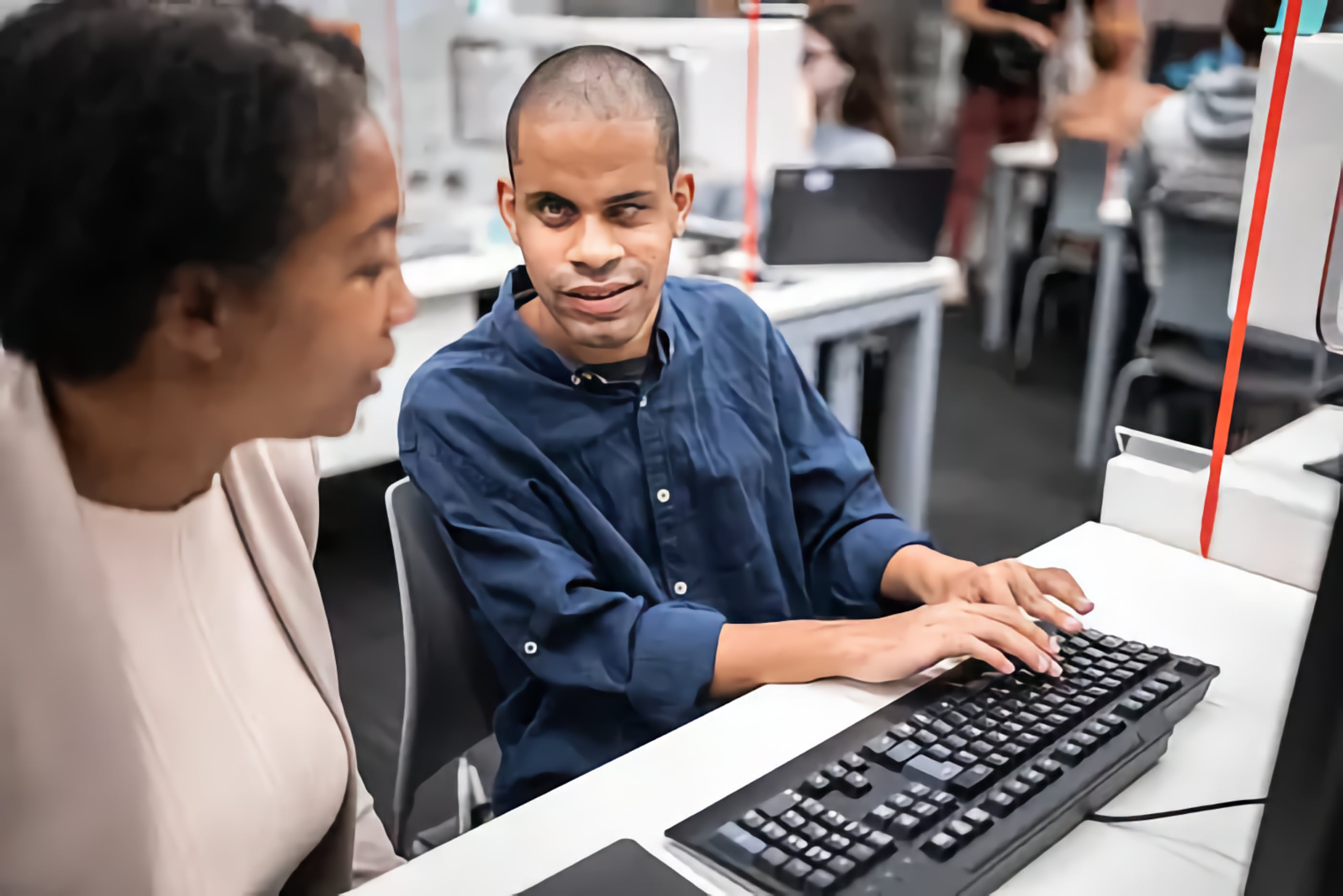 A Black woman and a Blind man with a Screen Reader collaborate, with the man typing on a computer keyboard