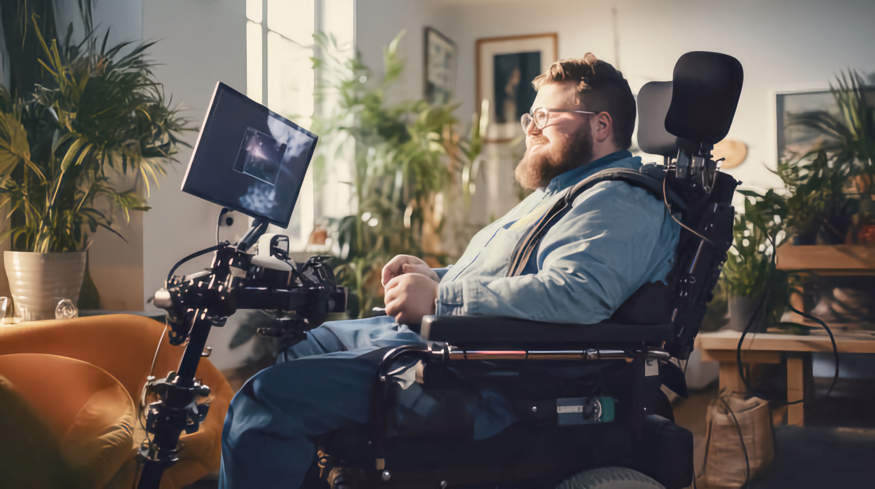 A White man with a beard sits in a motorized wheelchair inside a well-lit room filled with plants. He is smiling and looking at a mounted screen attached to his wheelchair.
