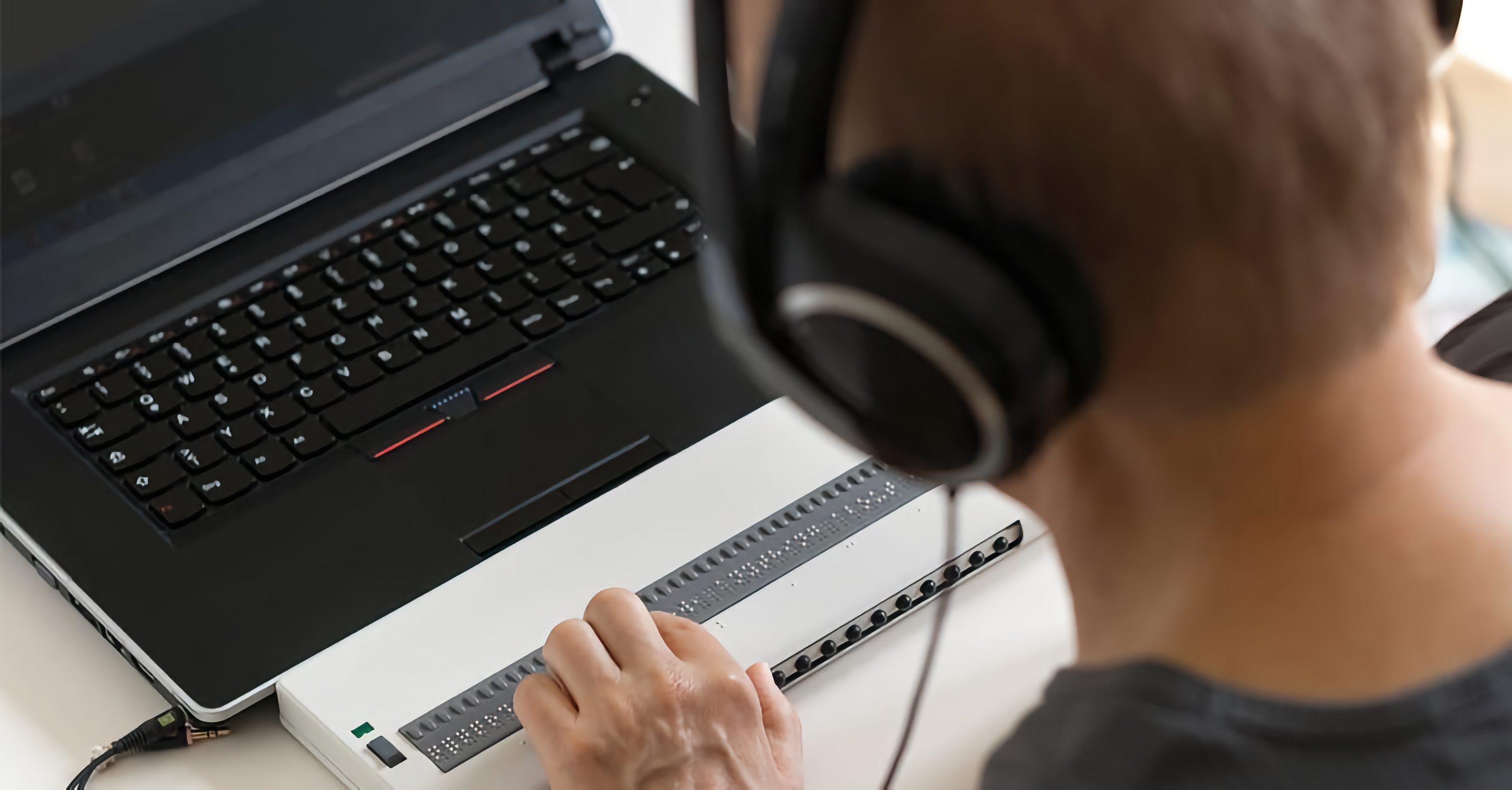 A blind man wears headphones and uses a refreshable Braille display and a screen reader while using the computer