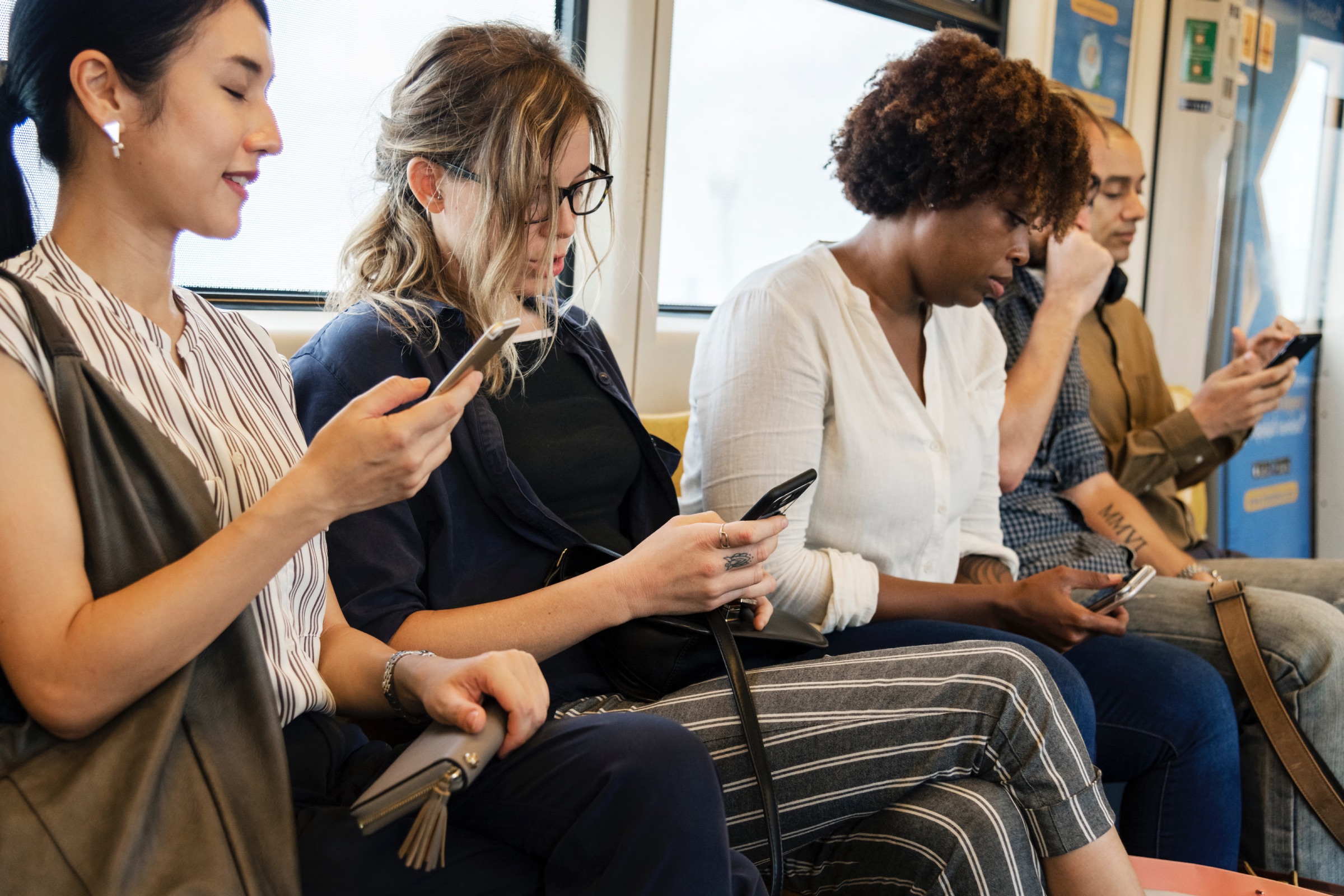 A diverse row of people riding public transportation, all looking at information on their mobile phones.