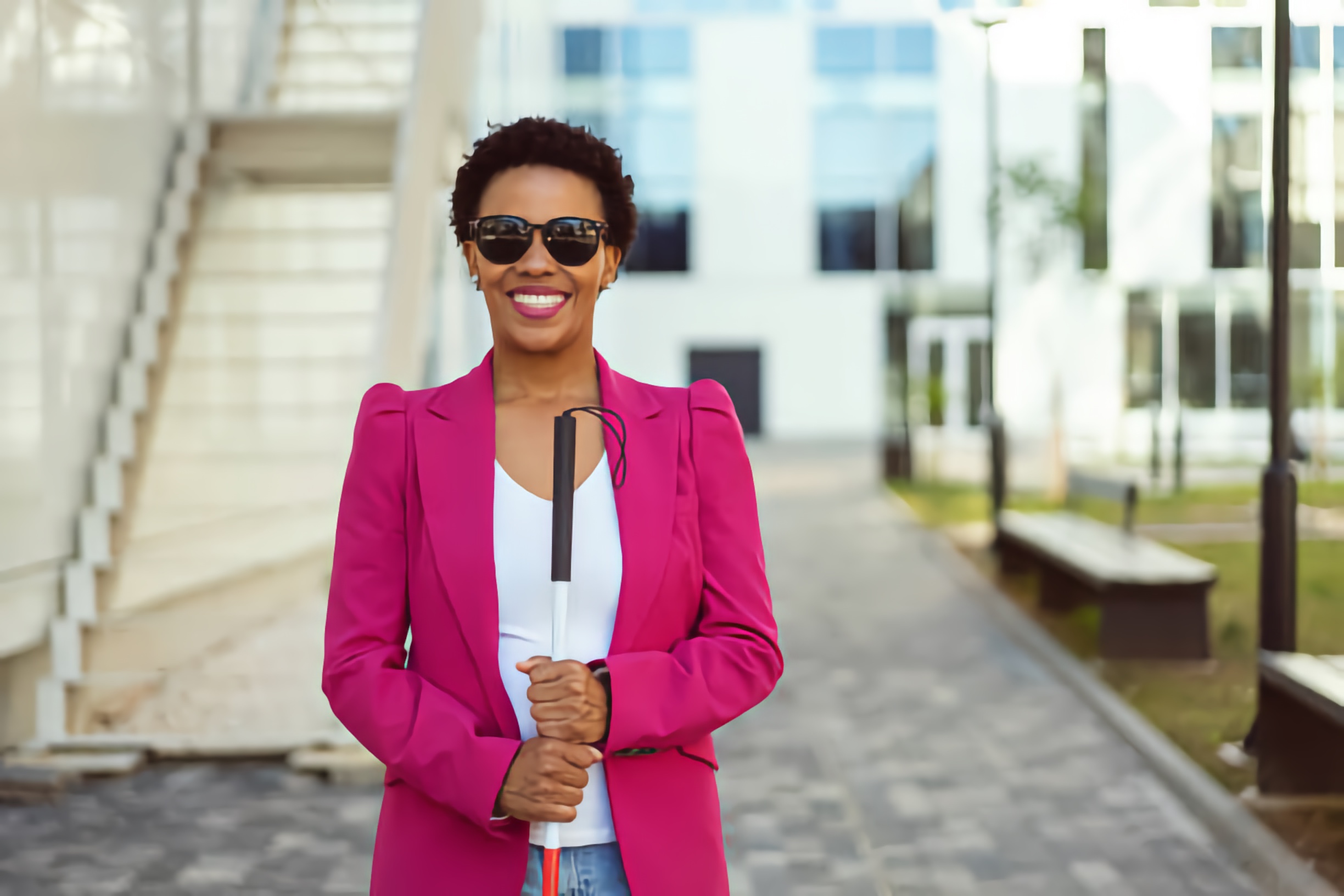 A smiling Black Blind woman wearing a pink blazer and sunglasses, holding a white cane in an urban setting