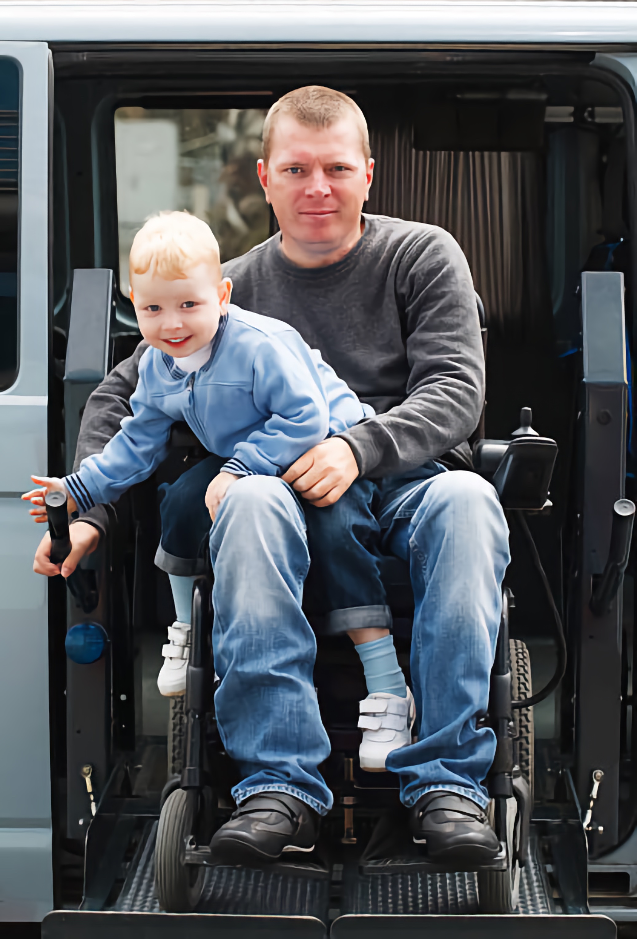 A smiling young boy sits on his dad's lap. The dad, using a powered wheelchair, exits a bus using the accessible ramp