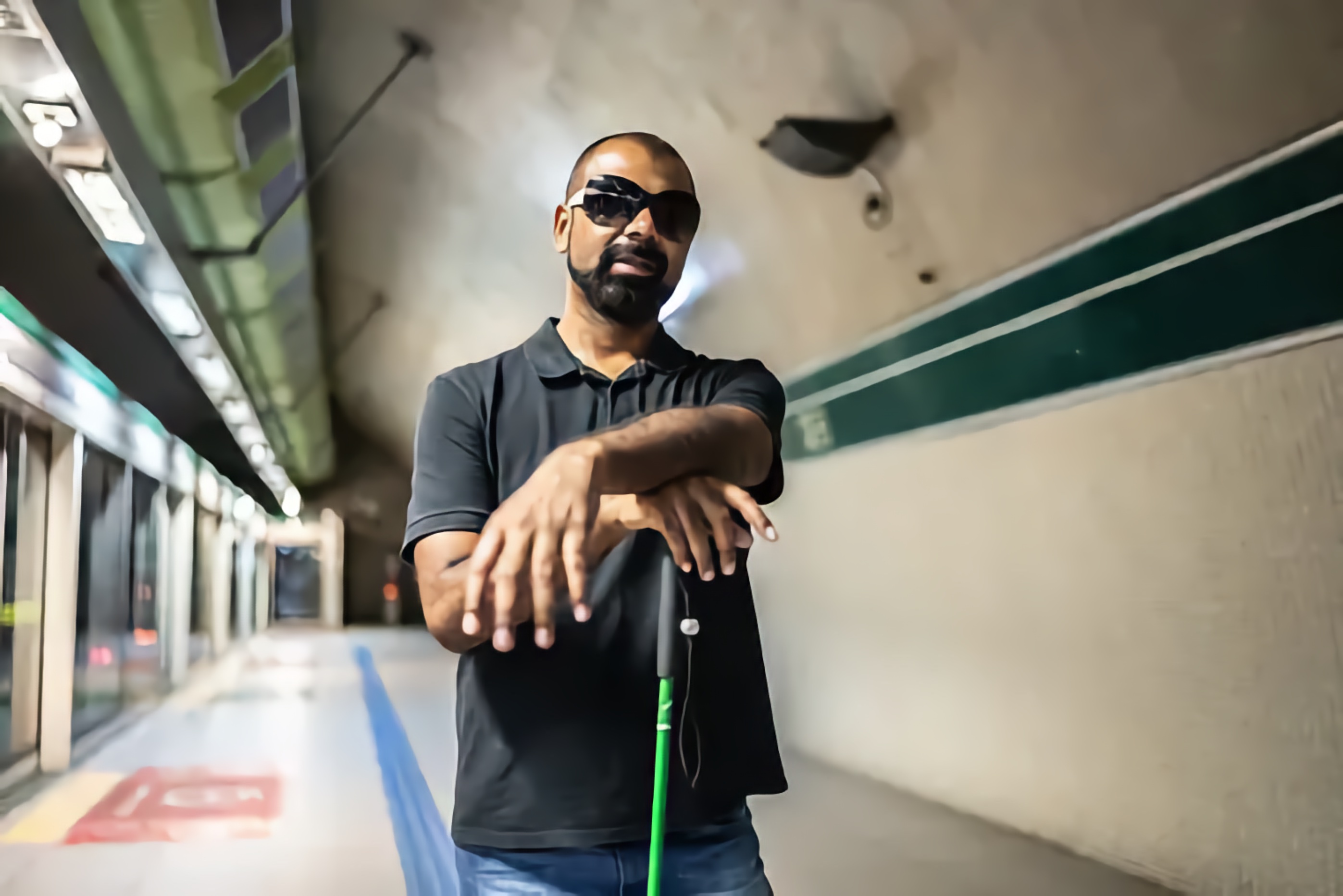 Blind Black man in dark grey polo shirt and sunglasses stands at a public transit station, arms folded over his cane, waiting for his ride