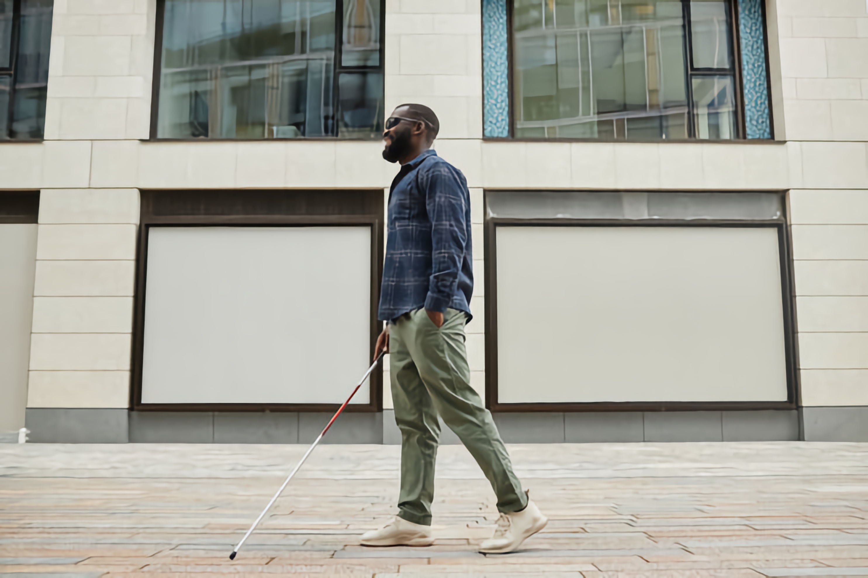 Blind Black man with a full beard, wearing a blue plaid shirt, walks along a sidewalk in a modern urban setting