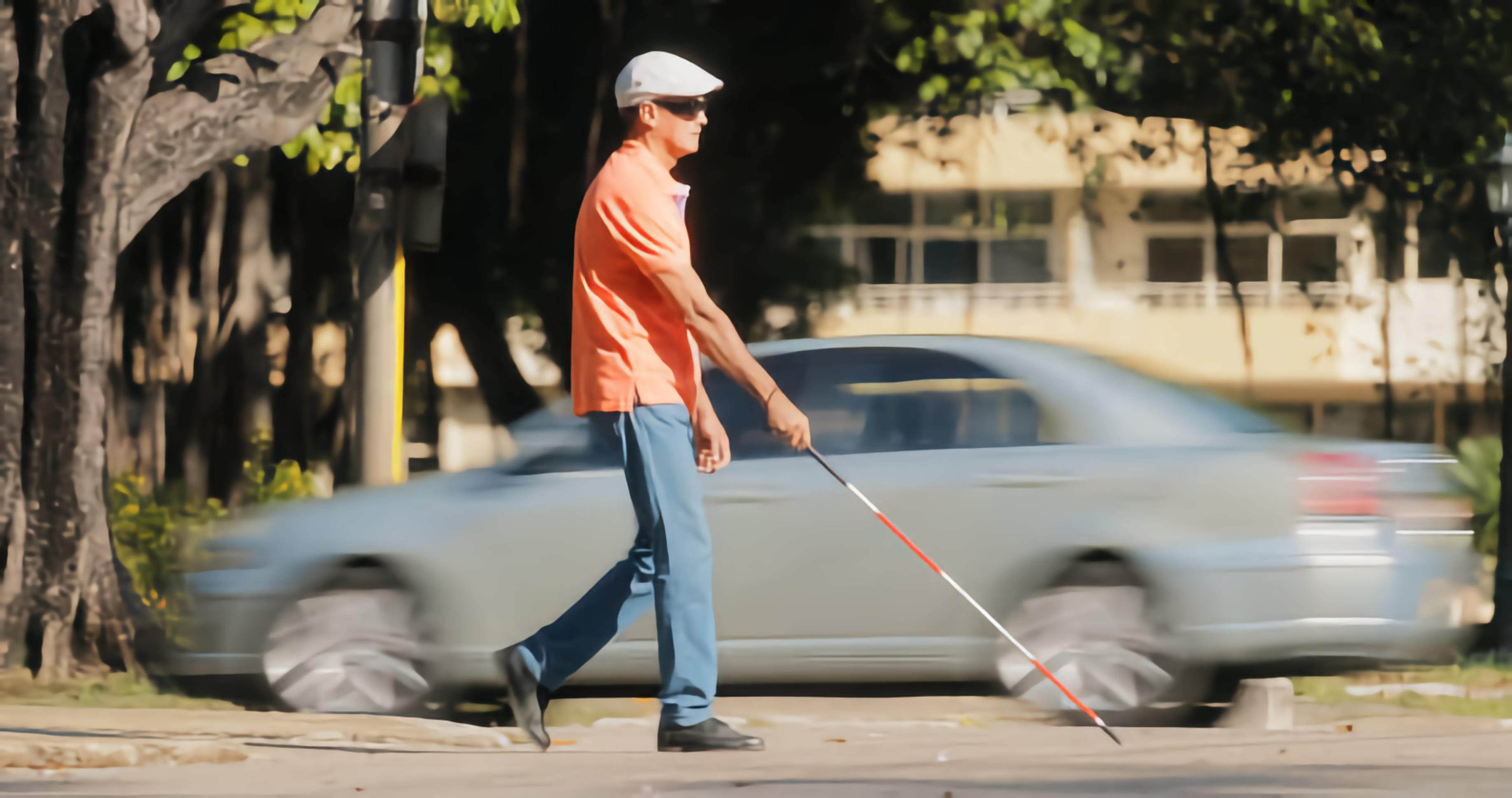 Blind white man wearing a peach-colored polo shirt and white cap walks along a sidewalk using his white cane while a blurred car whizzes past in the opposite direction