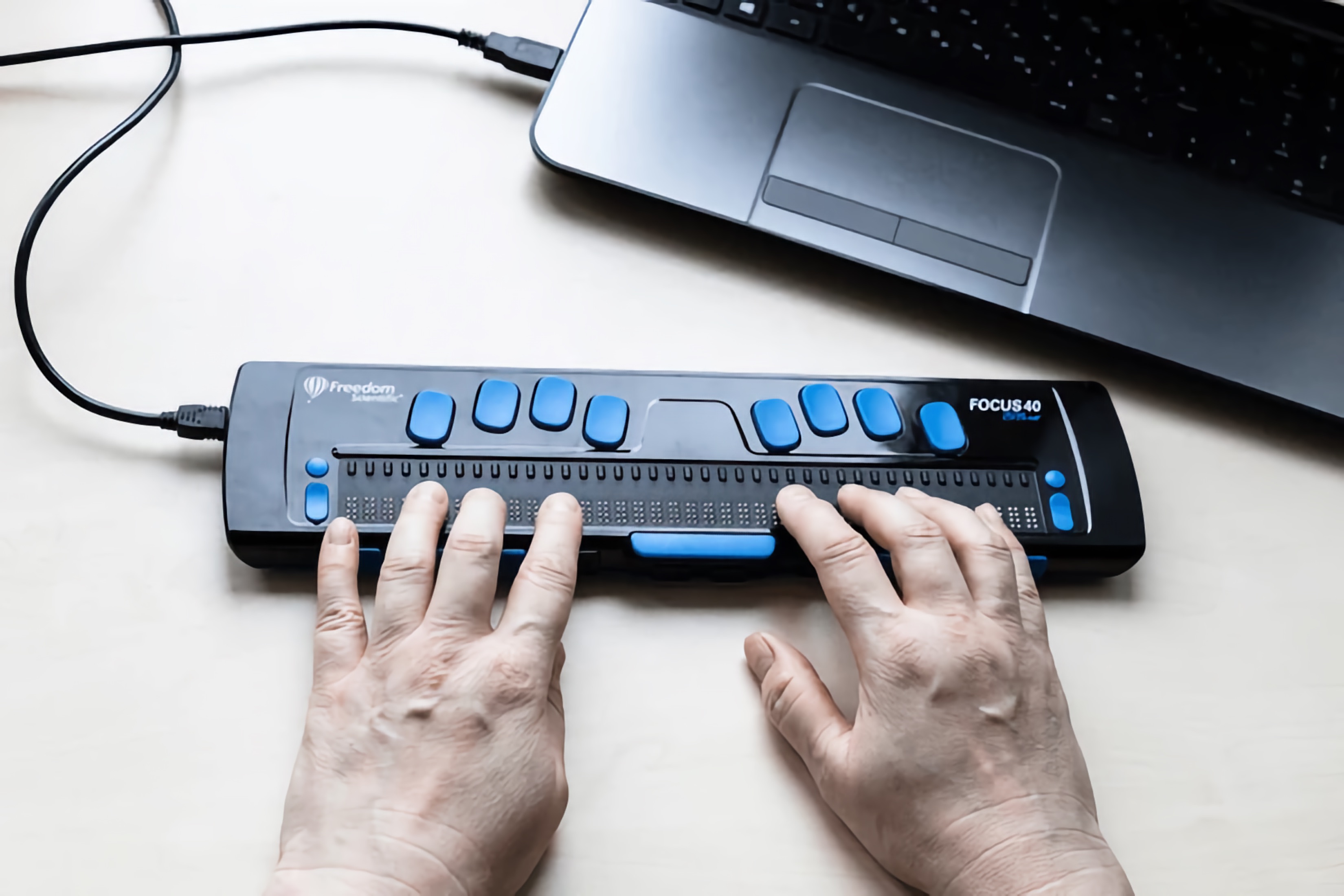 Close-up of hands using a Focus 40 refreshable Braille display connected to a laptop computer.