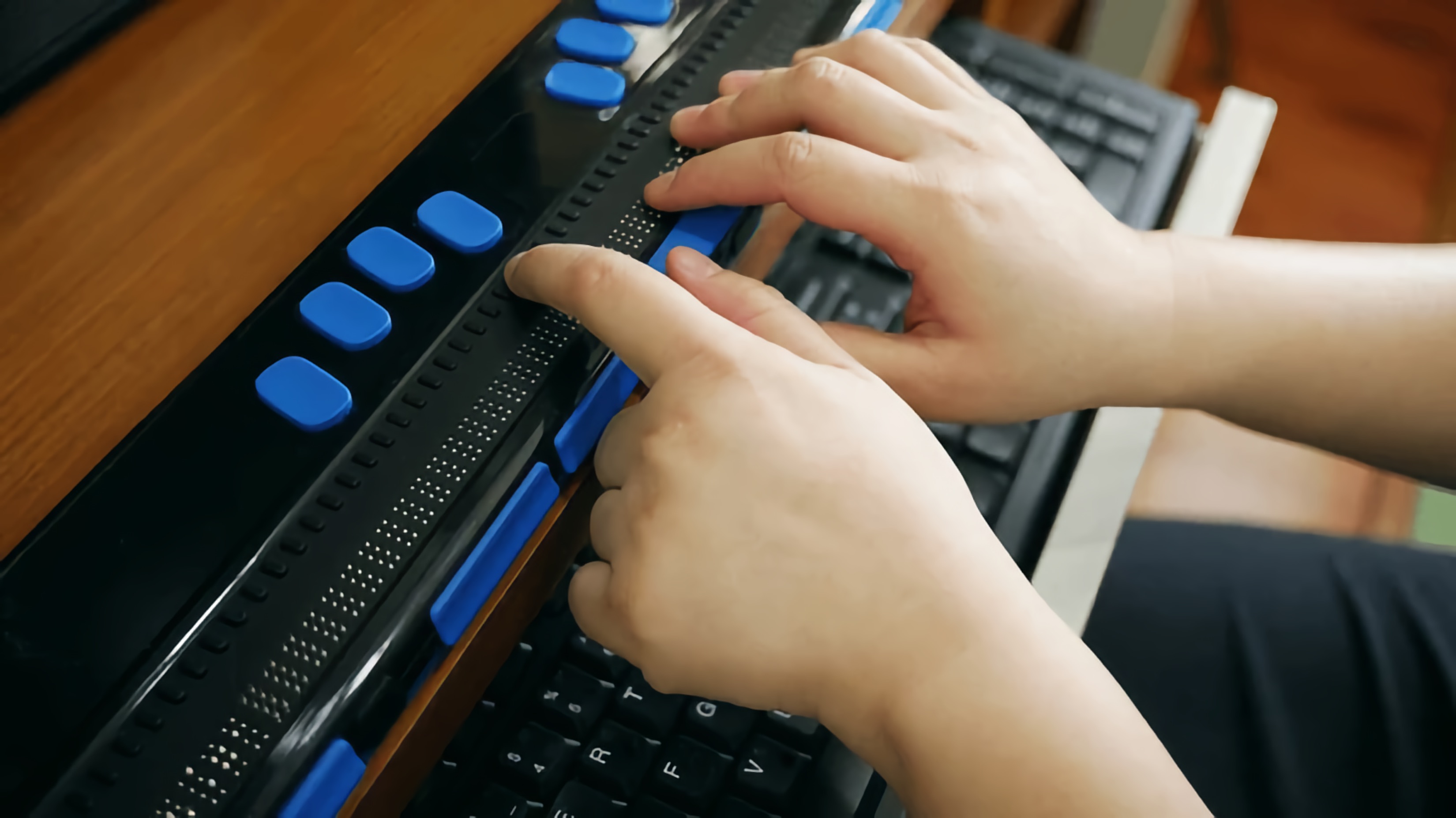 Close-up of hands using a refreshable Braille display connected to a computer.