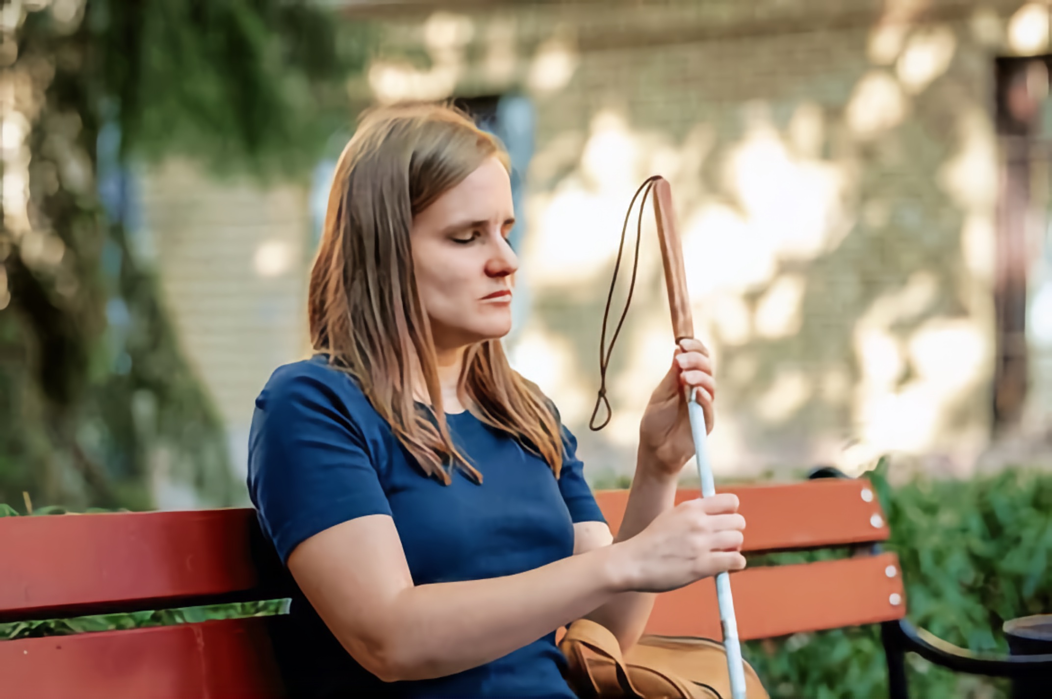 Youthful Blind woman with blond hair, seated at a bus stop bench, holds her white cane with her handbag beside her