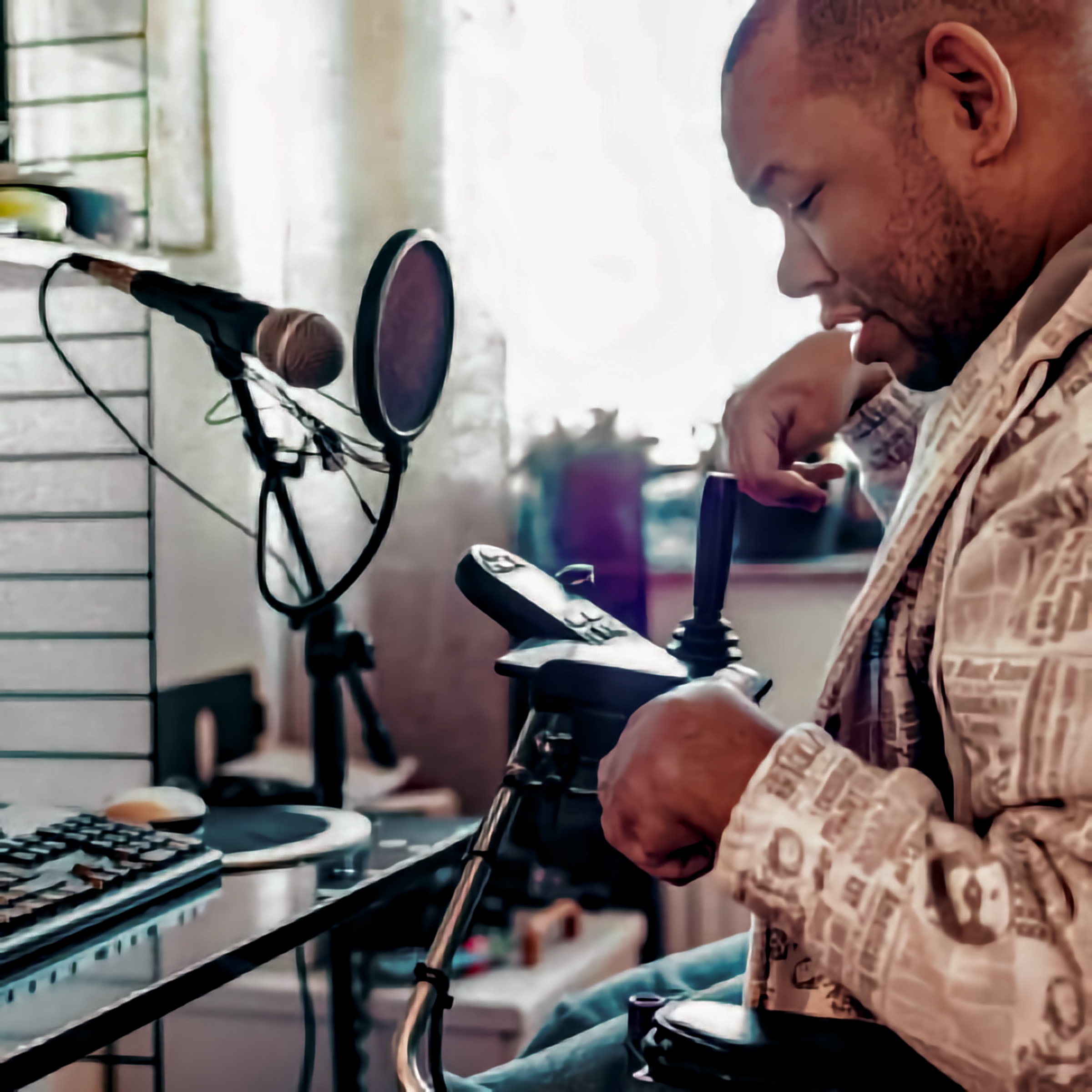 The image depicts a person using a powered wheelchair equipped with joystick controls, positioned at a workstation with adaptive technology, including a microphone and keyboard, enabling accessible communication and creative expression