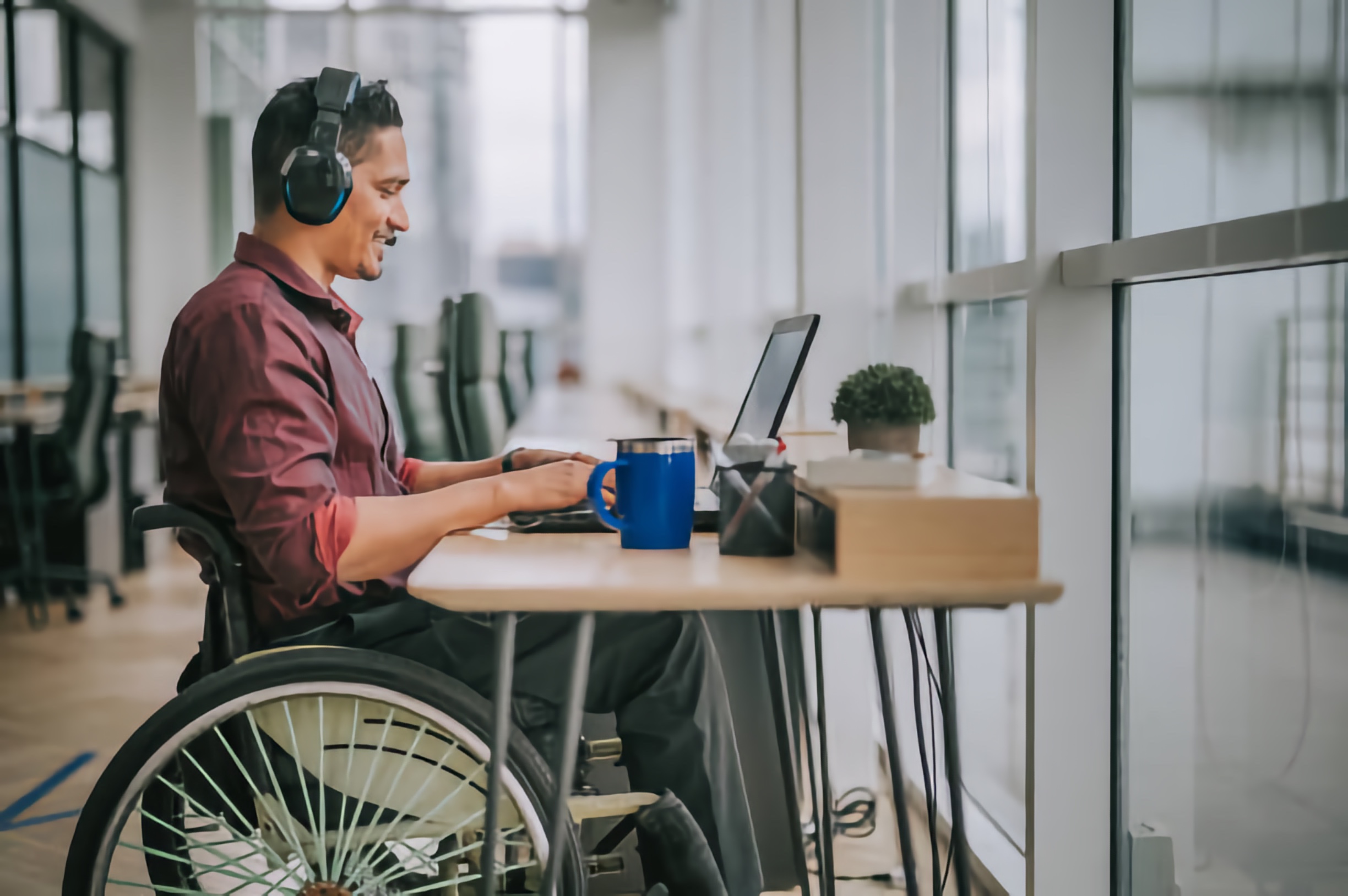 Man using a wheelchair, wearing headphones, smiling and working on a laptop at a desk in a bright office with a mug and potted plant.