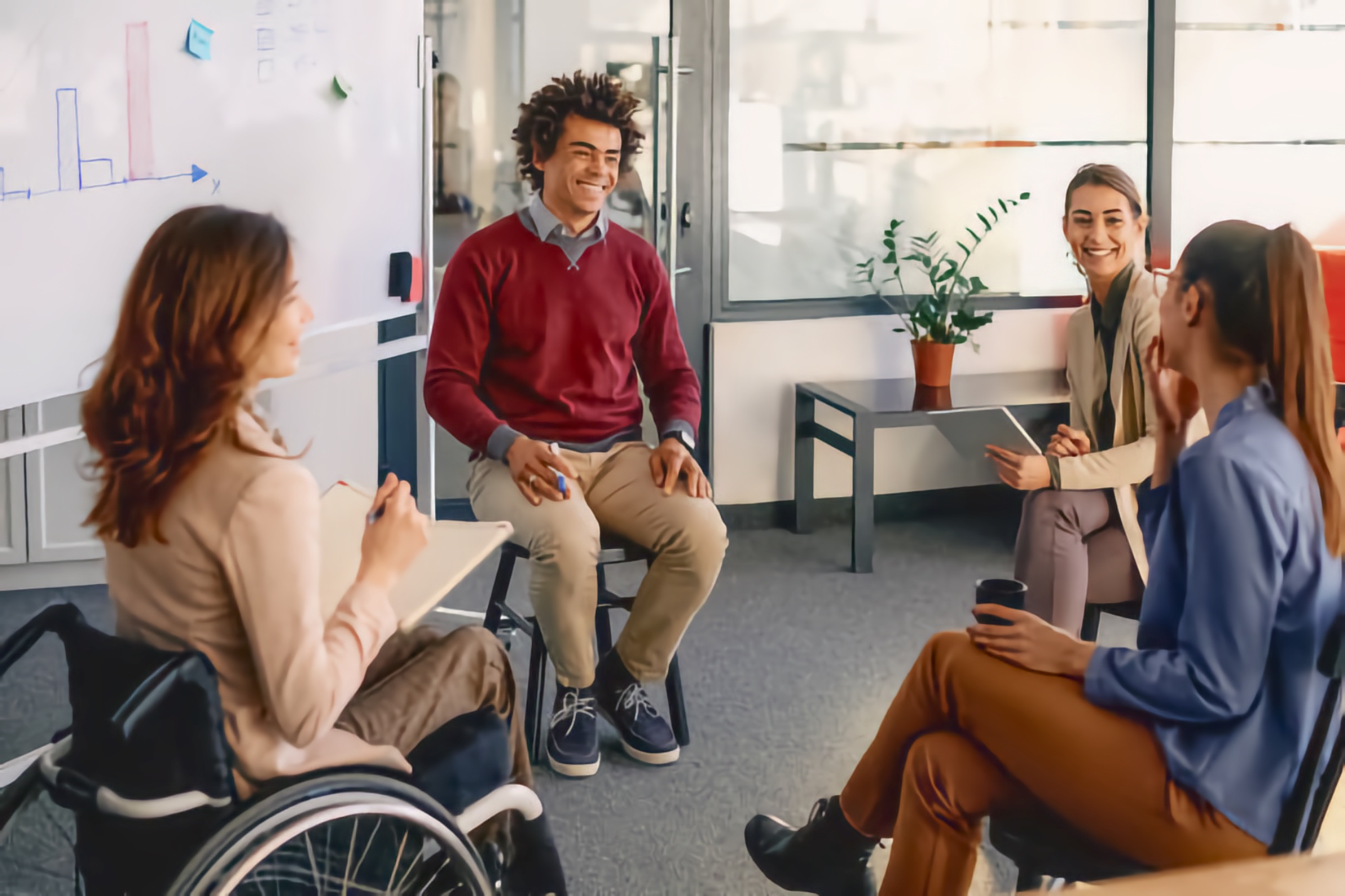A diverse group of professionals, including a woman using a wheelchair, sit in a circle smiling and engaged in conversation in a modern office near a whiteboard.