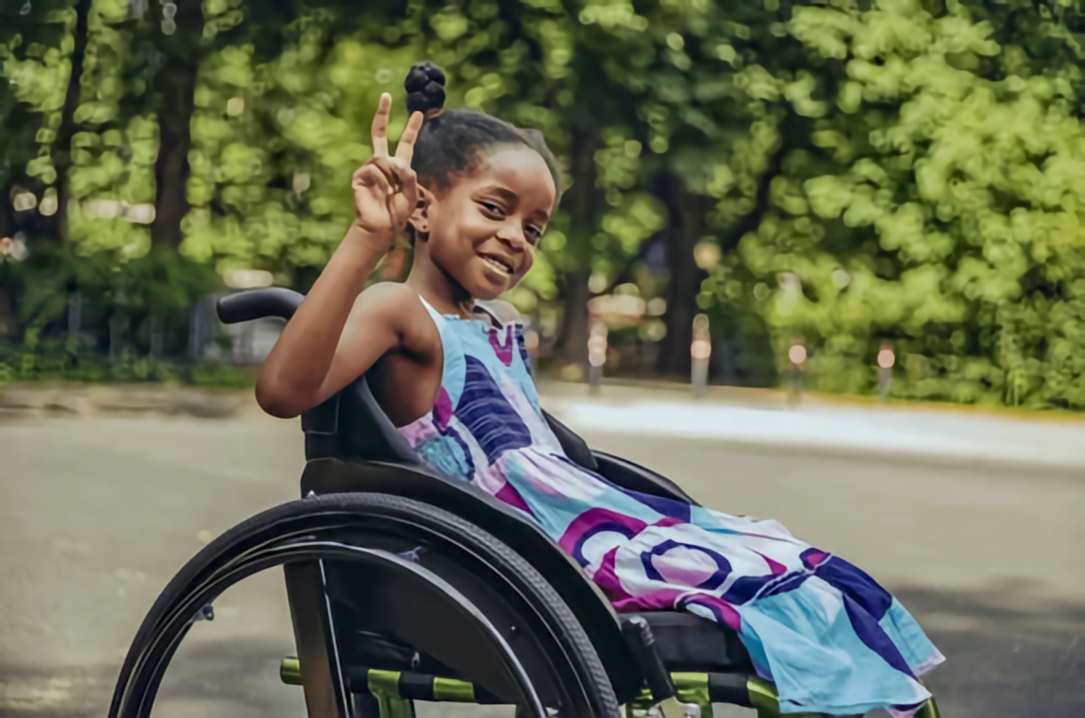 A Black girl uses a manual wheelchair and smiles as she holds up a peace sign. She wears a patterned summer dress and faces left on a paved road in a leafy park.
