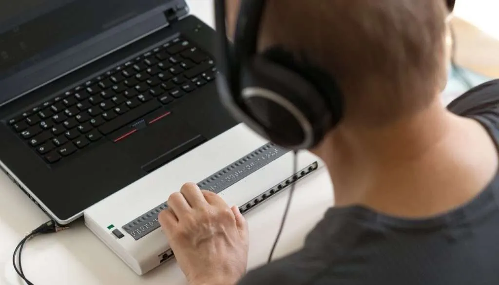 Rear view of a person wearing headphones reading a refreshable braille display connected to a laptop.