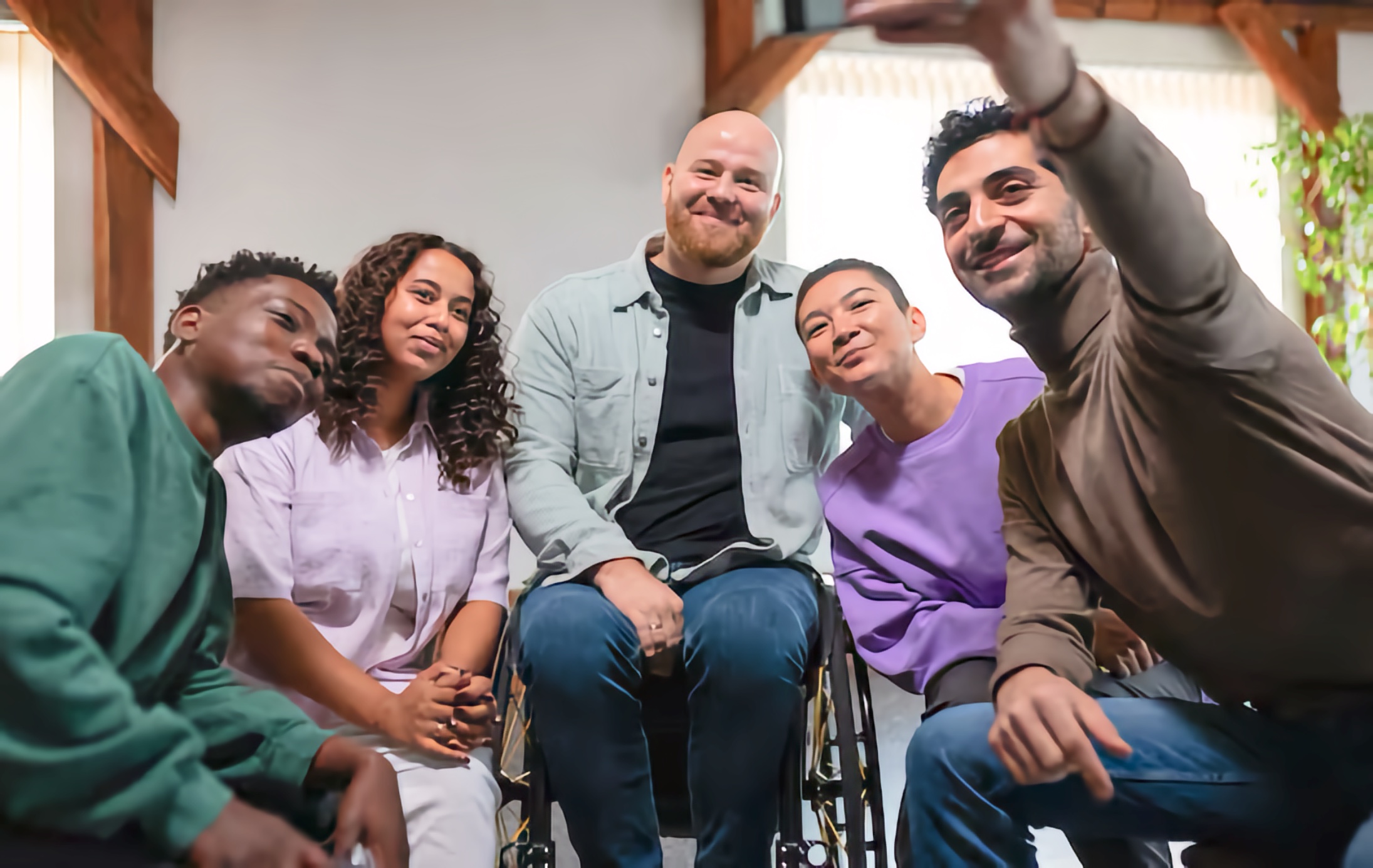 Five adults sit close, one in a wheelchair, as the person on the right extends a smartphone to frame the photo; wooden beams and a plant appear in the background.