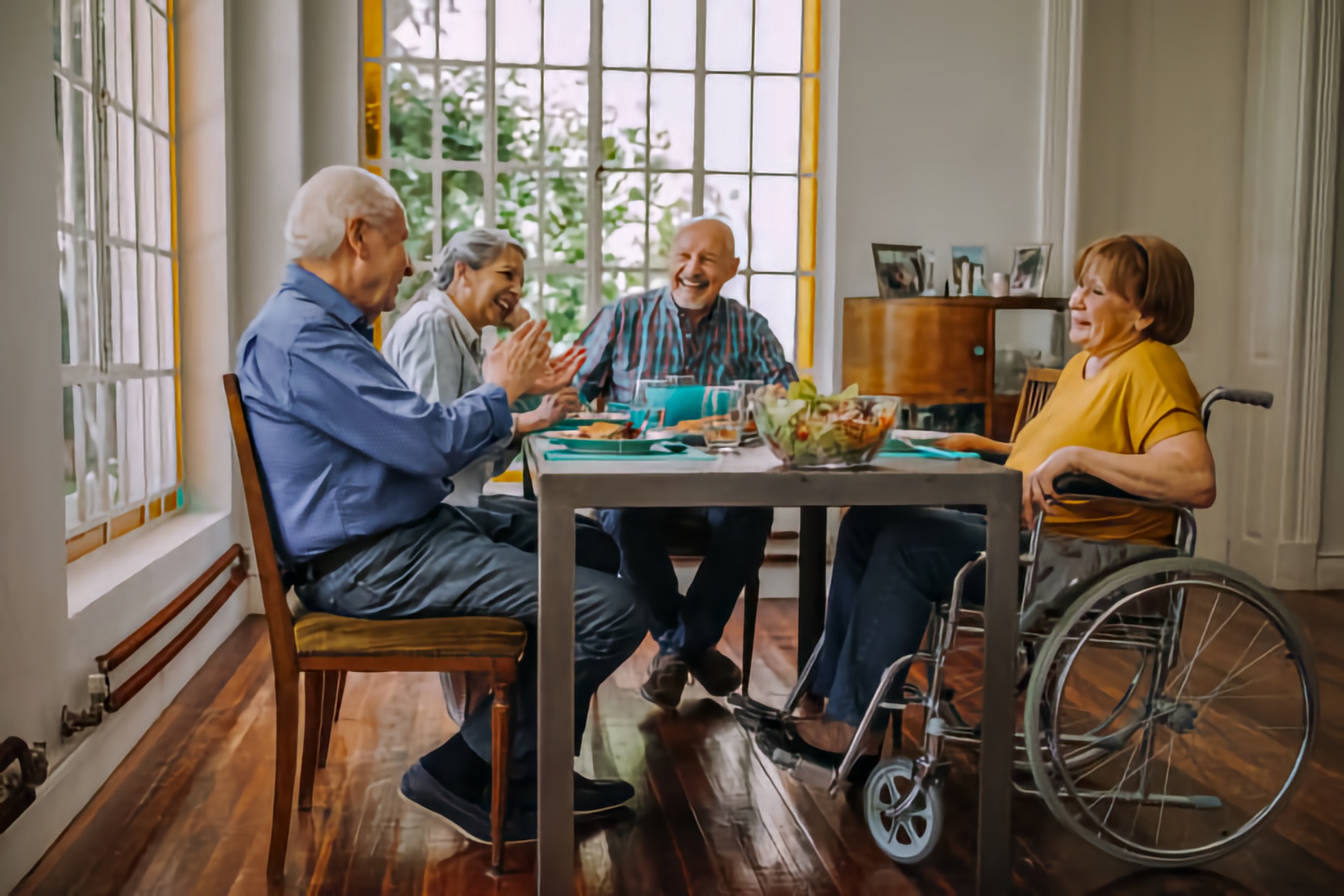 Four older adults sit around a dining table smiling, and one woman uses a wheelchair.