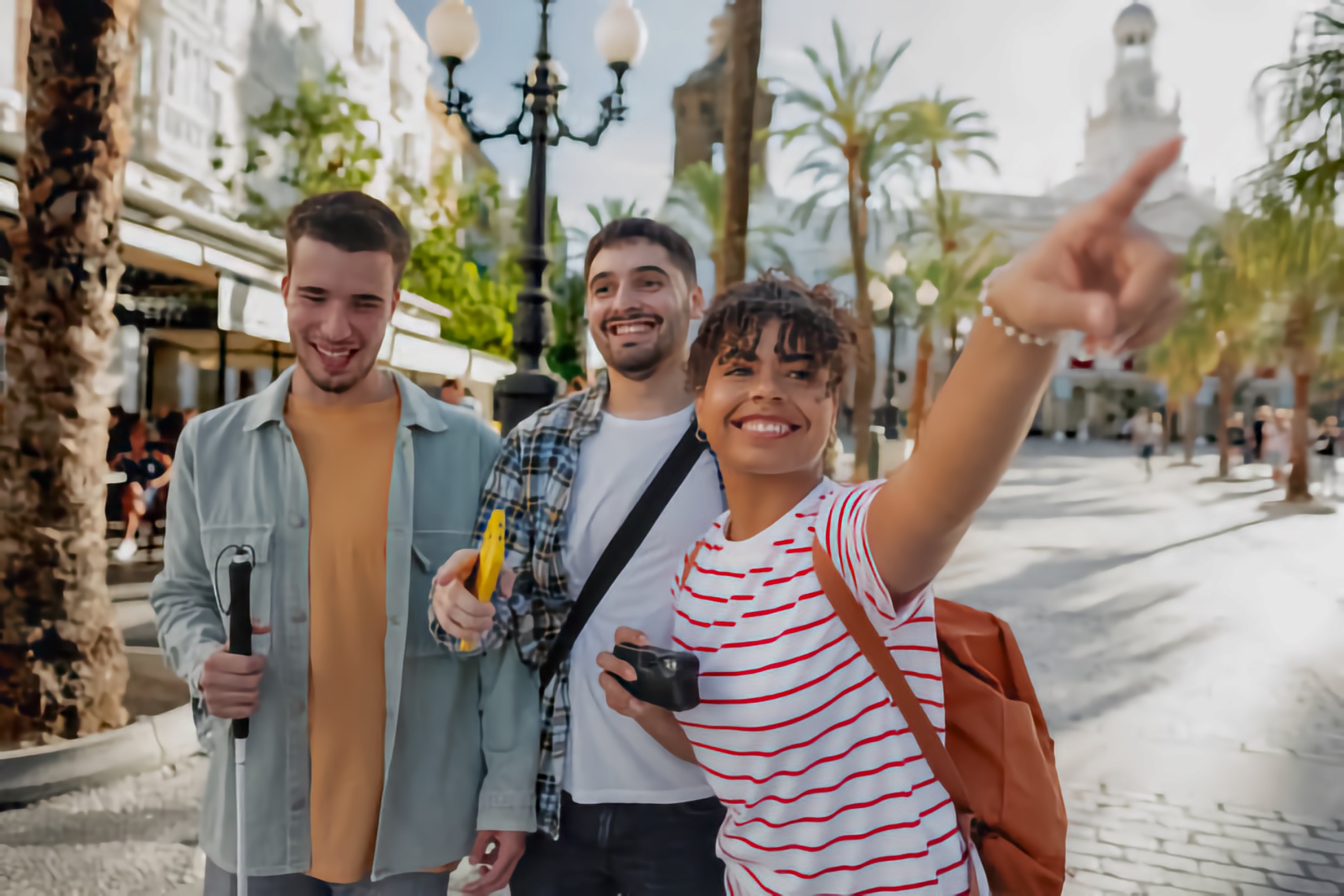 Three friends in a sunny plaza, one holding a white cane, another pointing ahead while the third smiles with a phone; cafés and palm trees line the walkway.