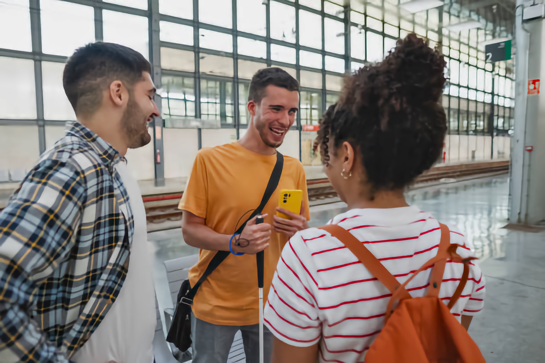 At an indoor rail station with a glass roof, a young man with a white cane shows something on a yellow smartphone to two companions wearing backpacks.