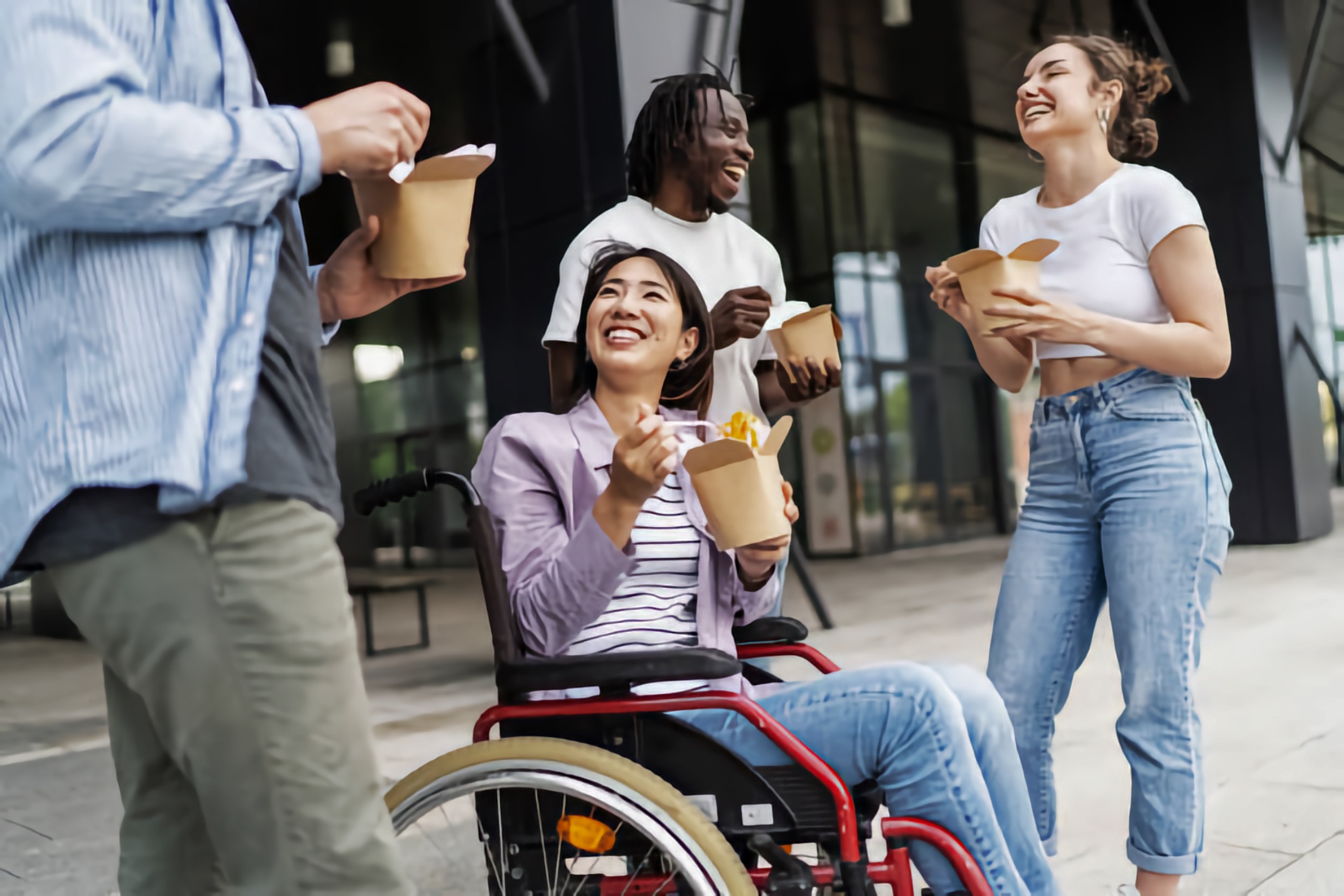 A young Asian woman using a wheelchair laughs with three friends, including a Black man and two other nondisabled adults, all eating takeout.