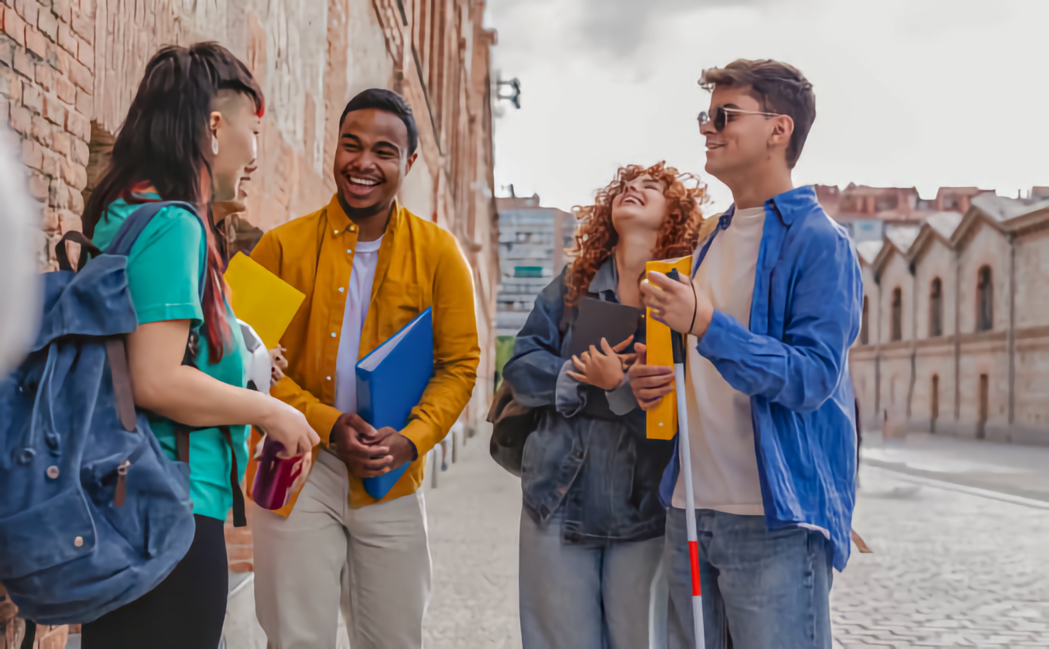 Four college-age friends stand chatting on a cobblestone street beside brick walls; one person with sunglasses holds a white cane while others carry notebooks and a backpack, all smiling.