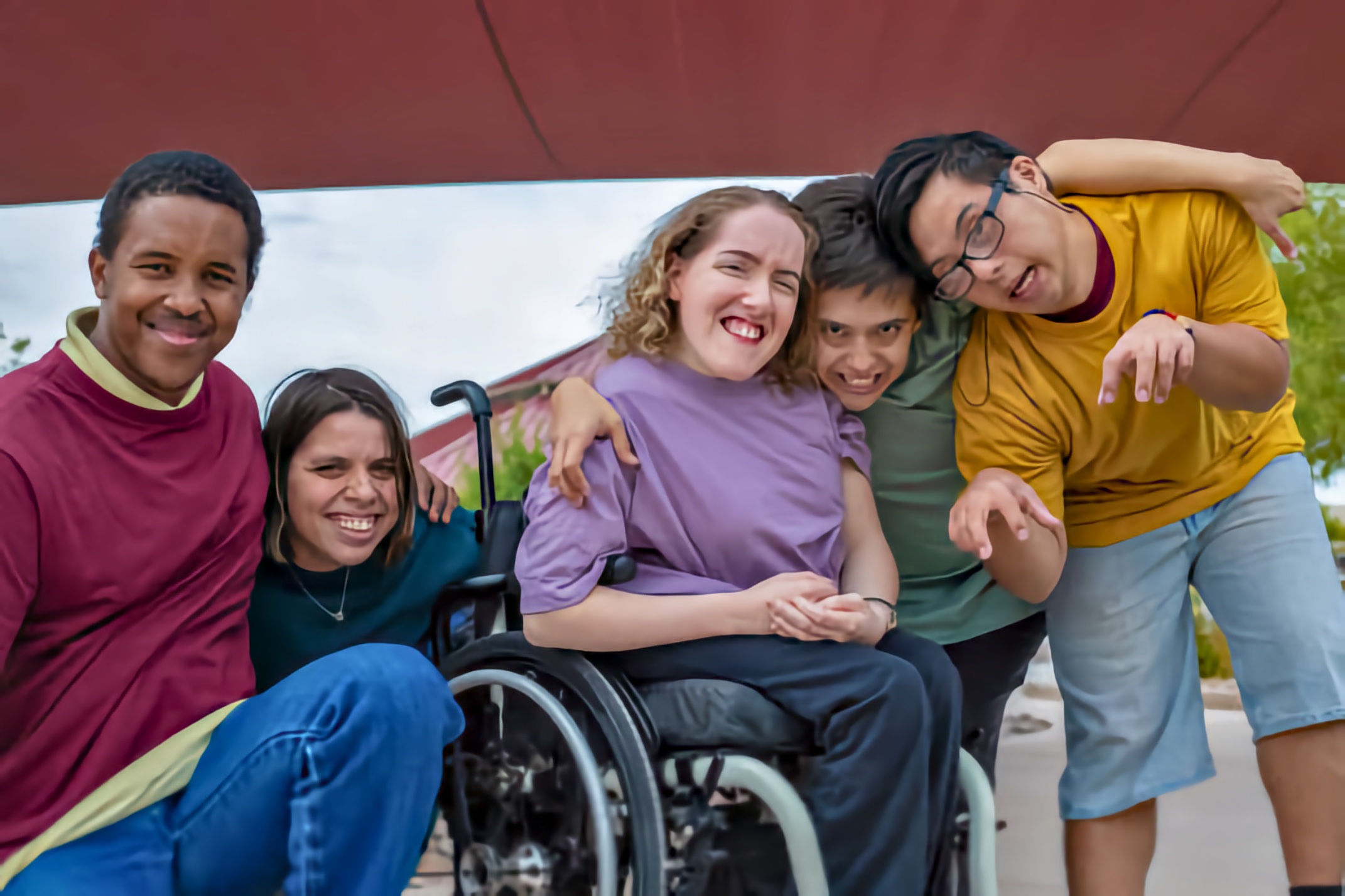 Five young friends huddle together outdoors and smile at the camera. A young woman who uses a manual wheelchair sits at the center while the others lean in with arms around one another under a red canopy.
