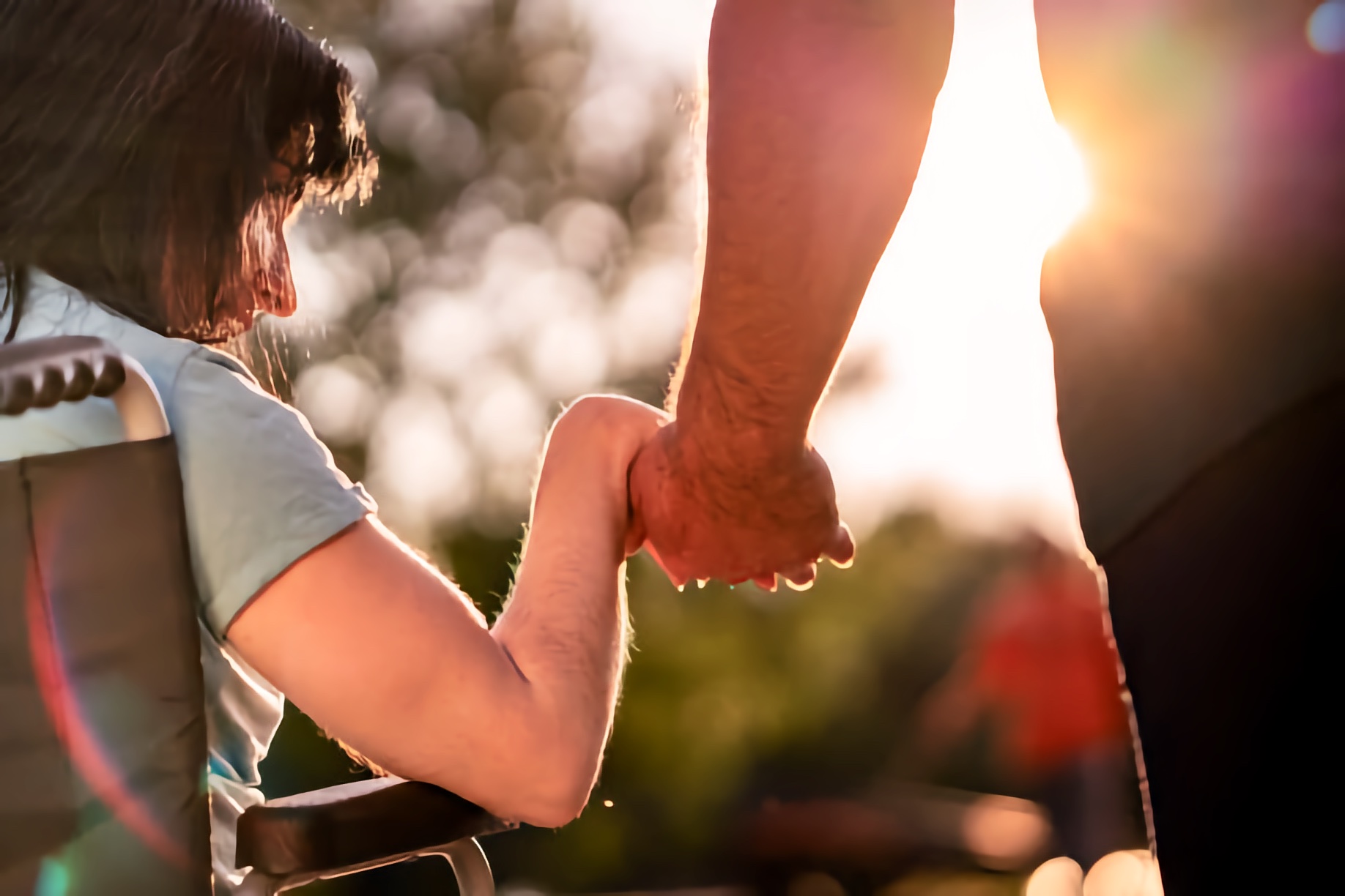 Close view of a wheelchair user’s left hand being gently held by a standing person while they move through a sunlit park; soft background with trees and warm light.