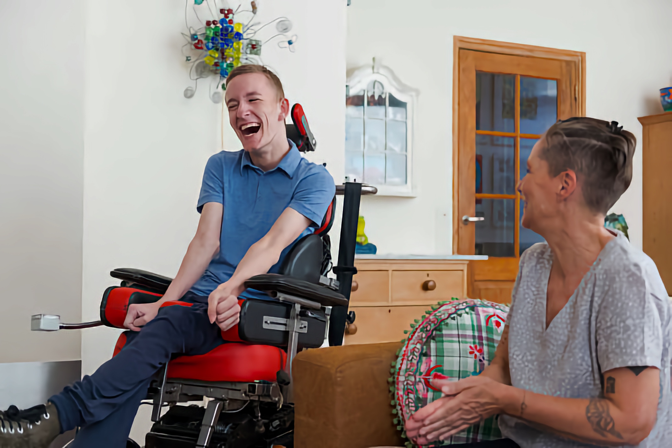 Young man in a power wheelchair laughs while chatting with a woman in a cozy living room.