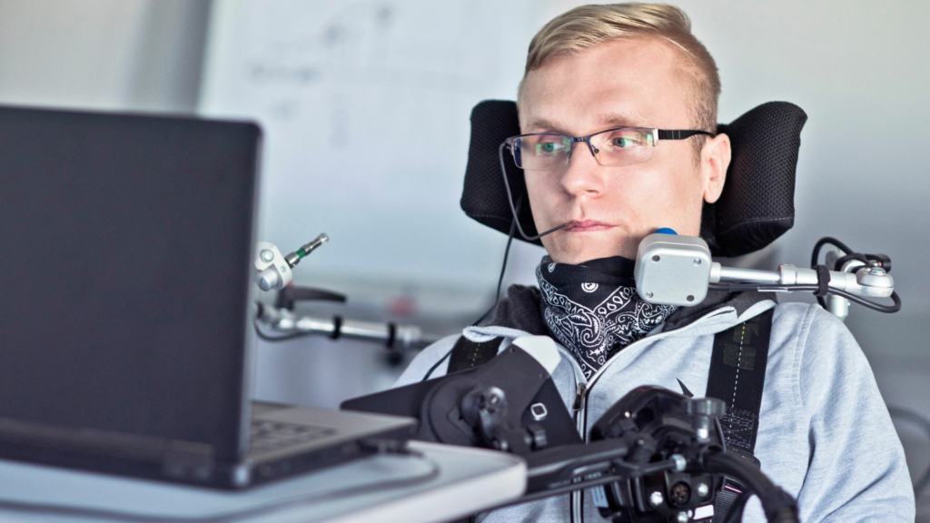 Young man in a powered wheelchair uses a mouth-controlled joystick while looking at a laptop on a desk.