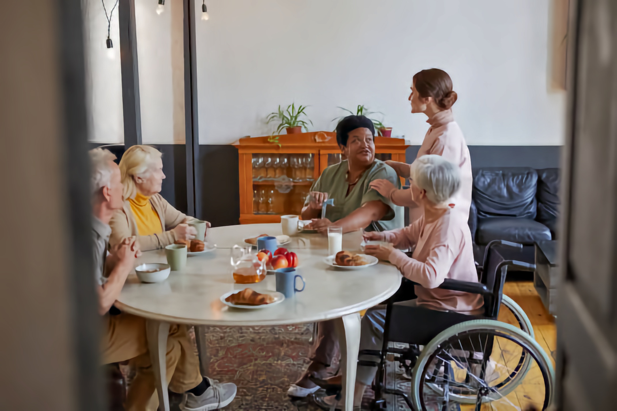 An older white woman in a wheelchair sits at a table with an older white man, an older white woman, and a middle-aged Black man, while a younger white woman stands beside them.