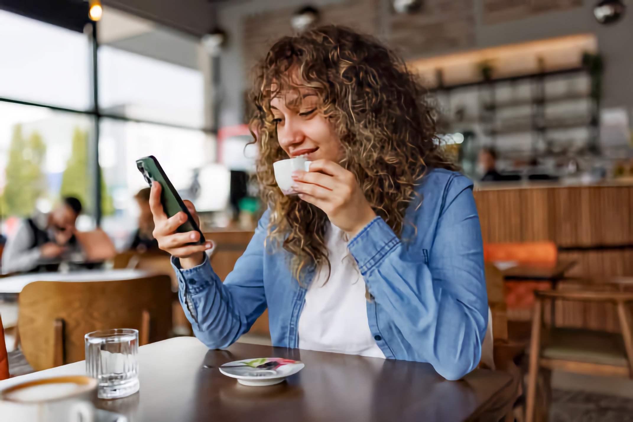 Smiling woman holds a cup of coffee while looking at her smartphone in a cozy café.