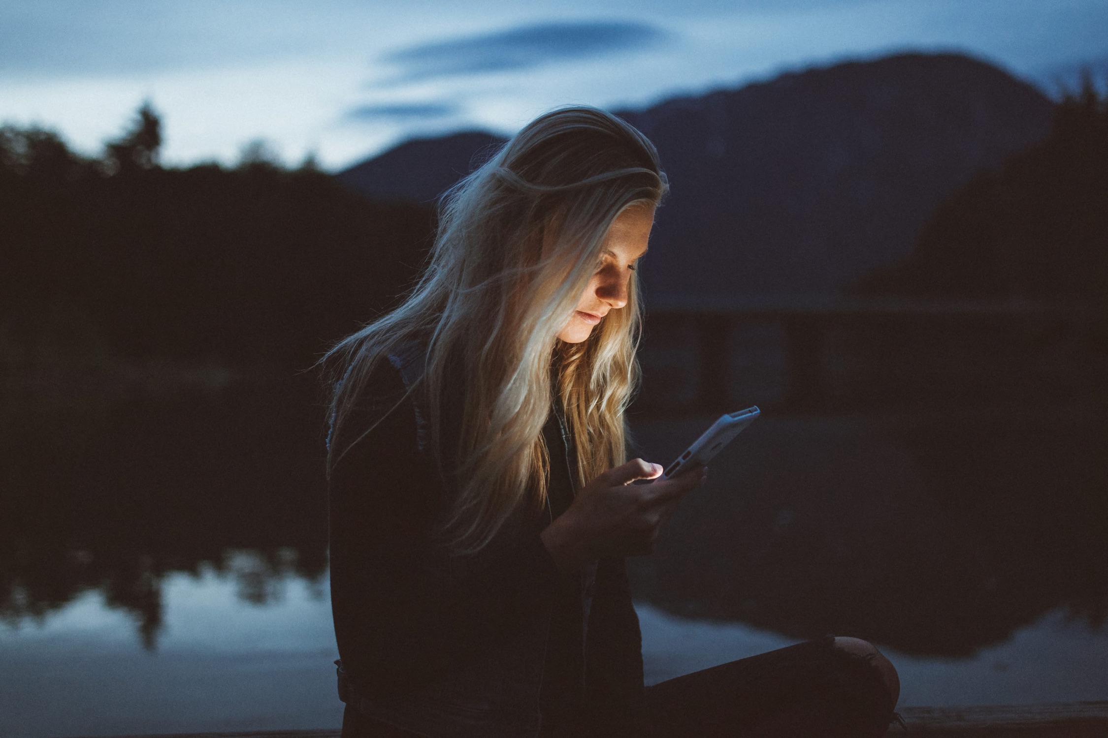 Woman sits by a lake at dusk, her face softly lit by the glow of a smartphone screen.