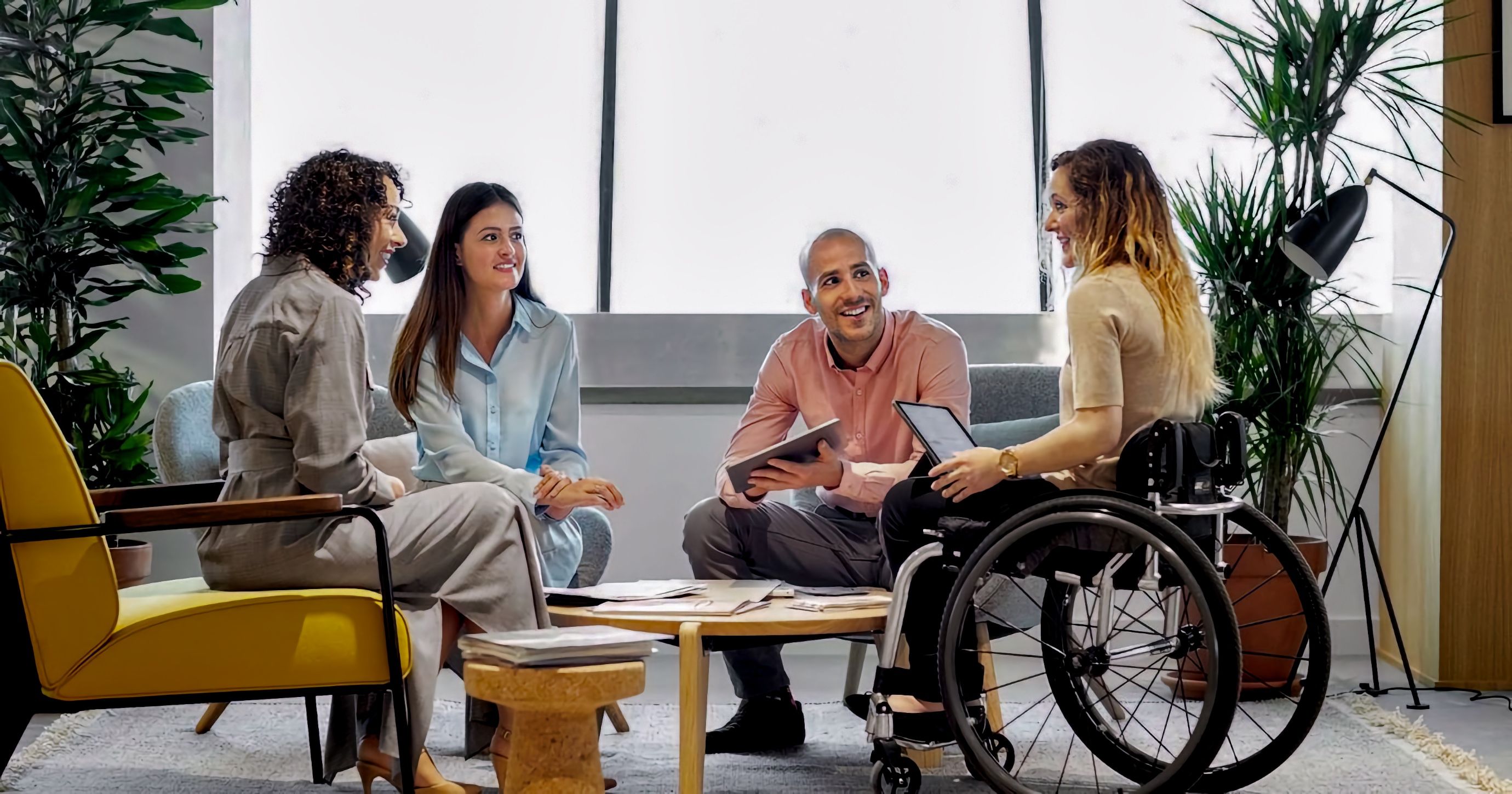 A mixed-race team meets in a lounge area. A white woman using a manual wheelchair speaks while holding a tablet, and three colleagues—two women and one man—sit on a sofa and chair facing her and listening.