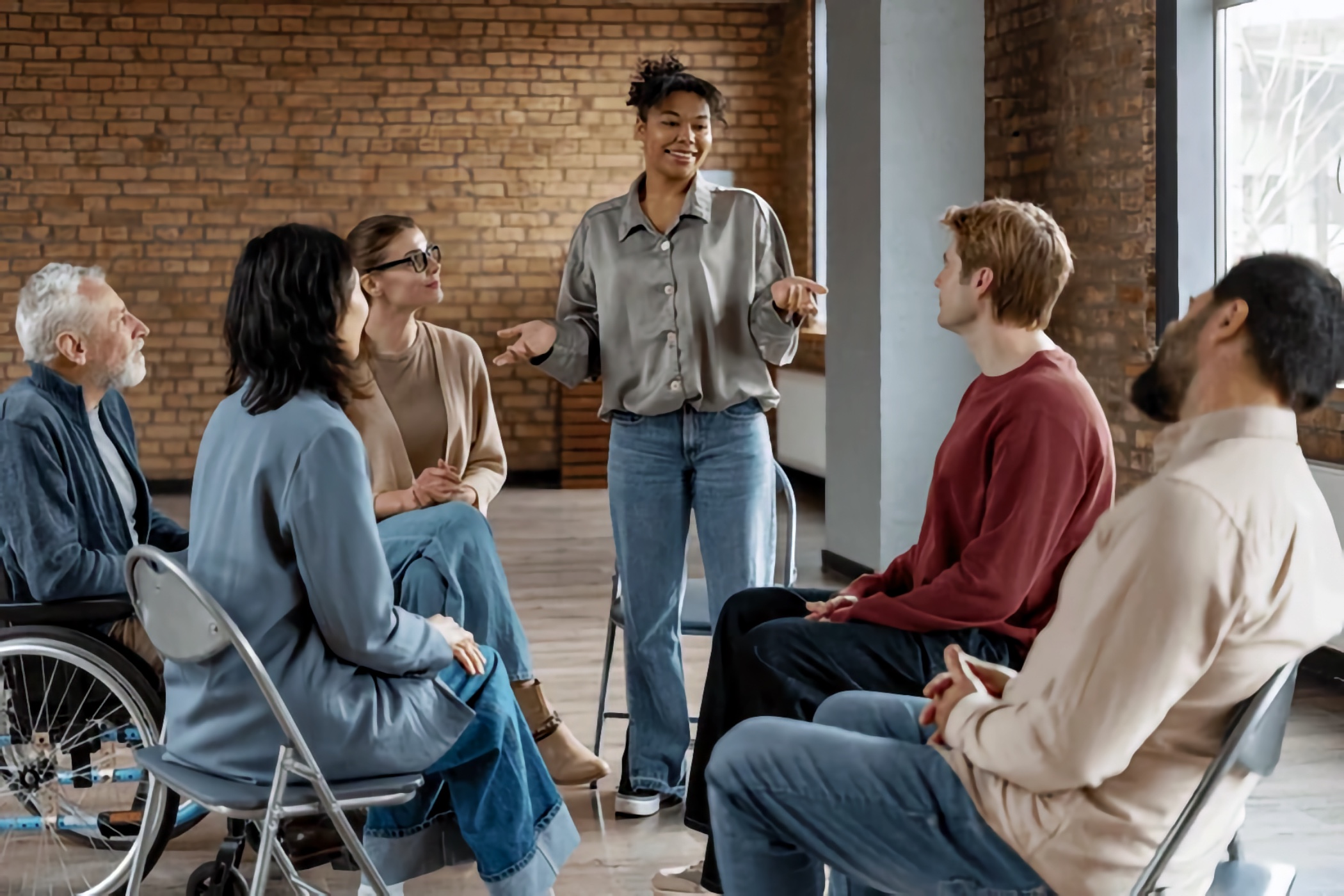 A Black woman stands and leads a discussion in a brick-walled room. Six adults sit in a circle facing her; on the left, an older white man uses a manual wheelchair.
