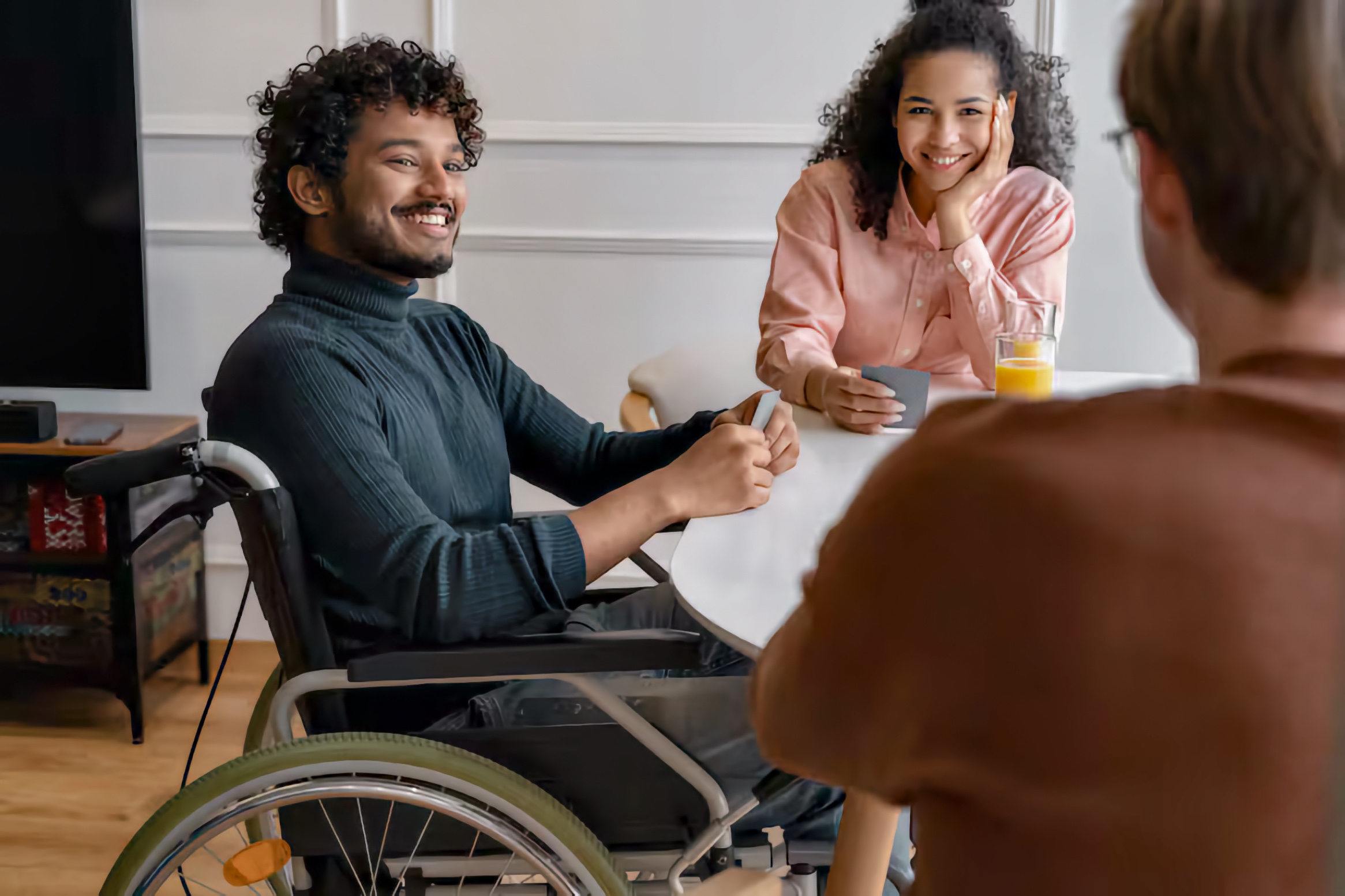 Mixed-race group of three friends at a round table; a Black young man using a manual wheelchair smiles while holding playing cards, a woman smiles with her chin in hand beside a glass of orange juice, and a third friend sits with their back to the camera.