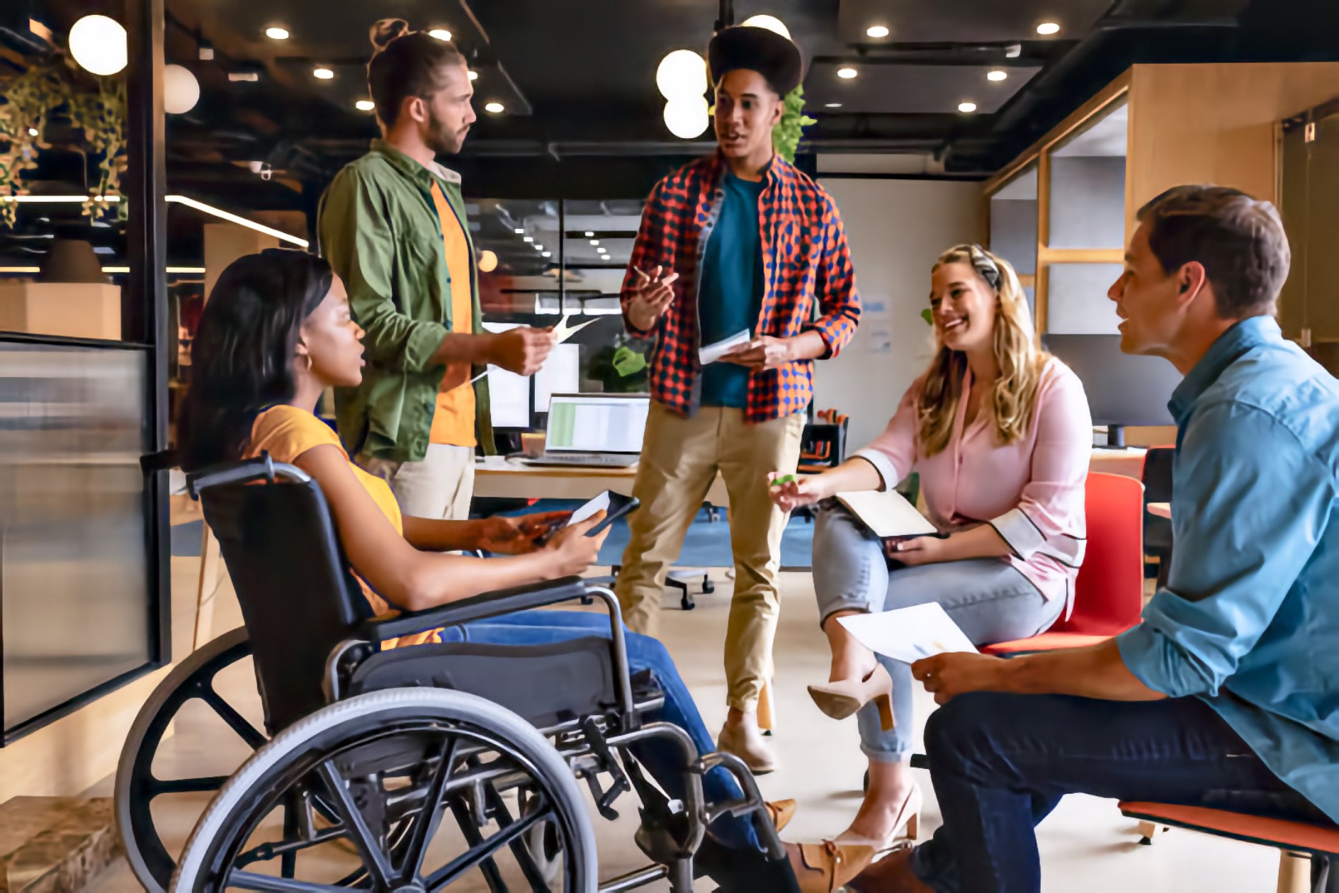 Five young adults meet in a shared work space for a group discussion. A young adult Black woman who uses a manual wheelchair sits at the left holding papers and speaking to the group. Two young adult men stand nearby holding notes—one white man with a beard and one Black man with a high bun who gestures while talking. Two young adult white colleagues sit facing them, listening and holding papers. The scene shows collaborative planning in an accessible environment.