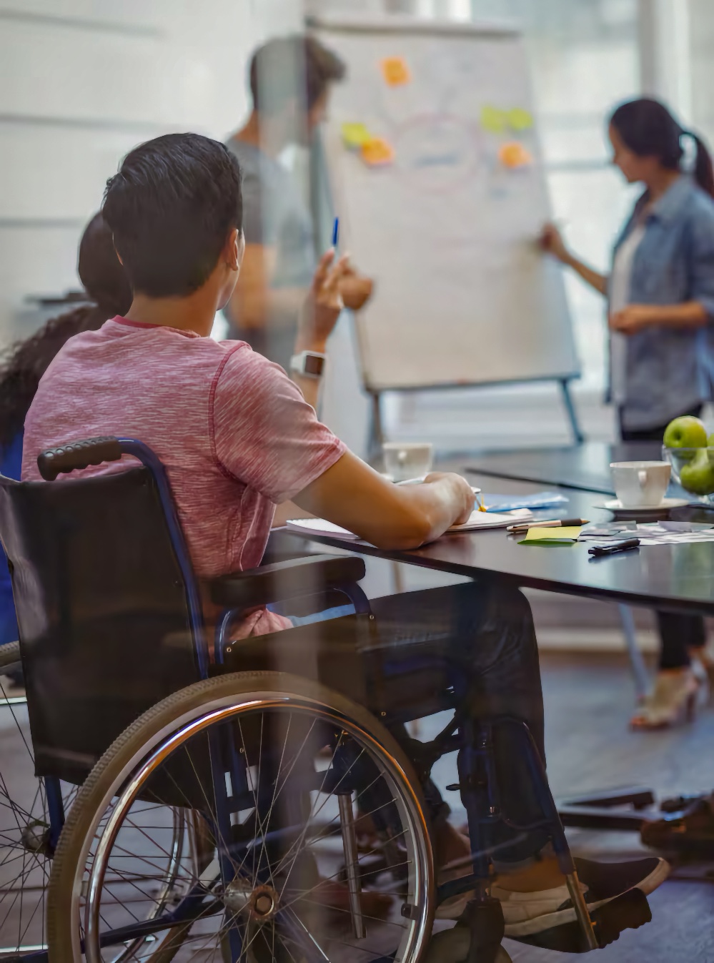 In a bright community meeting room, a young adult man using a manual wheelchair sits at a table and raises a blue marker toward a flip chart covered with colorful sticky notes. In the background, a young adult man stands near the flip chart, and a young adult woman with her hair in a ponytail points toward the notes as if presenting, while notebooks, pens, coffee cups, and green apples are spread across the table.