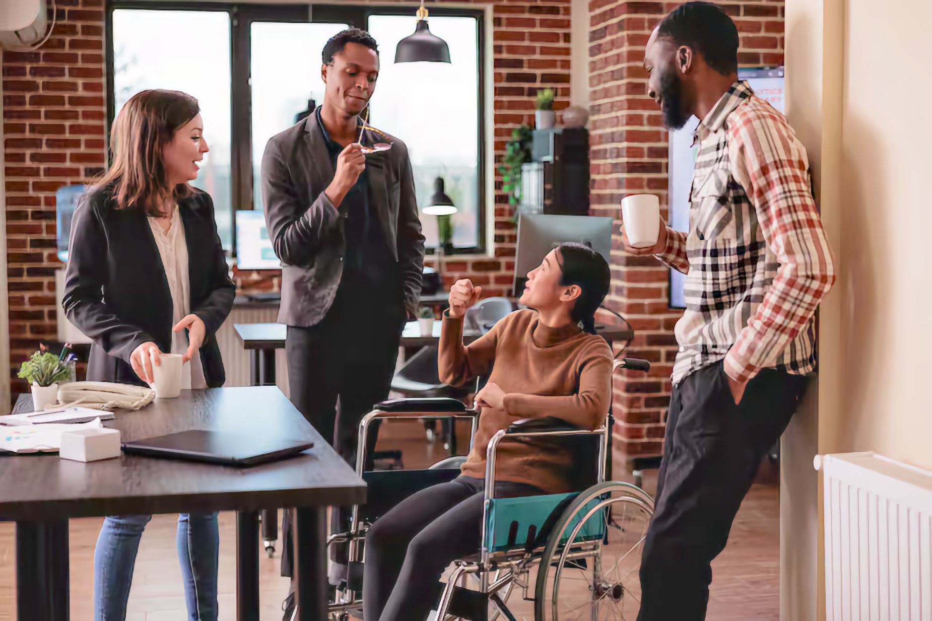 In a brick-walled community gathering space, an Asian young adult woman using a manual wheelchair laughs and gestures as she talks with three standing friends. A White young adult woman holds a mug beside a table, while two Black young adult men stand nearby—one holding his glasses near his mouth and the other holding a mug—as the group chats together.