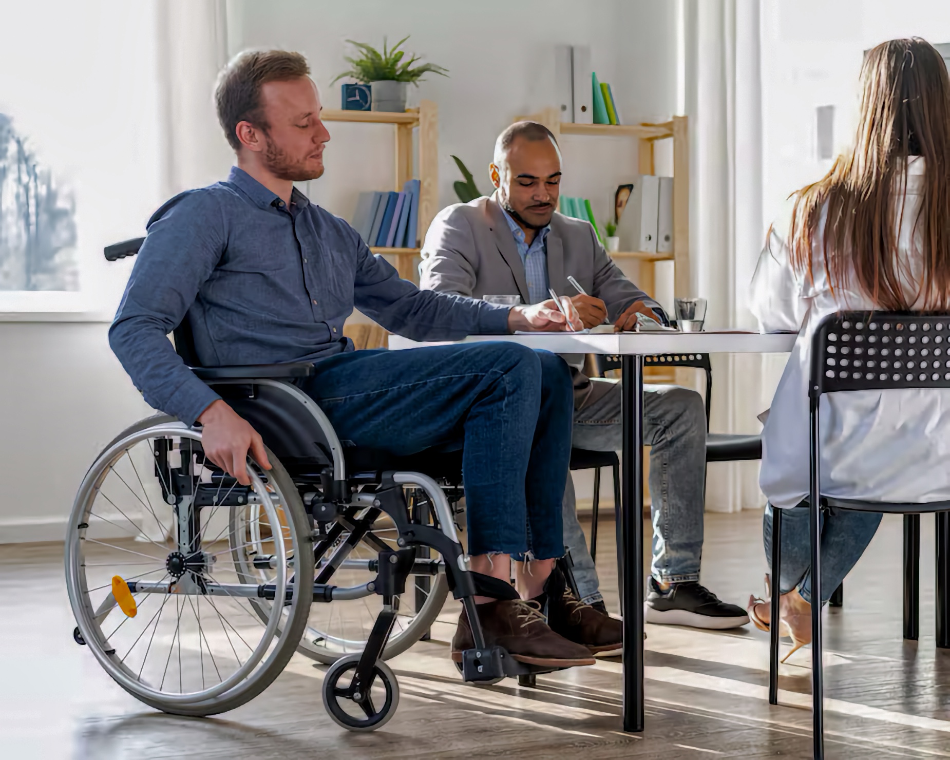 At a small community planning meetup in a bright shared room, a White adult man using a manual wheelchair sits at a round table with two other adults. A Latino adult man in a gray blazer writes notes on paper while listening, and a White adult woman with long brown hair sits with her back to the camera, facing the group as they work together on community event plans.