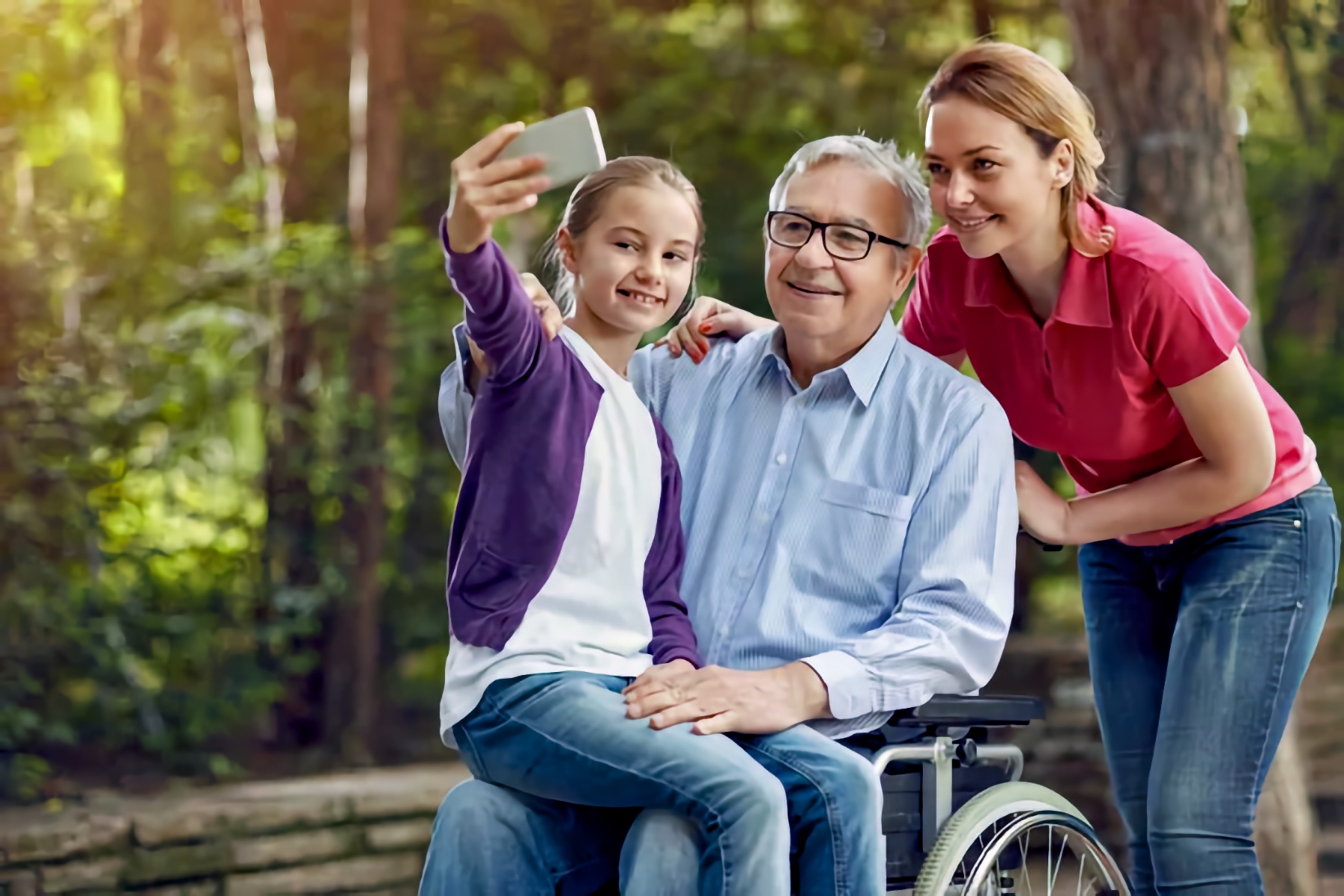 In a sunny park with trees in the background, a young girl holds a smartphone at arm’s length to take a selfie with her grandfather and her mother. The older adult man, wearing glasses and a light blue button-down shirt, sits in his wheelchair and smiles while the girl sits on his lap. An adult woman in a pink polo shirt leans in close from the side, smiling toward the phone.
