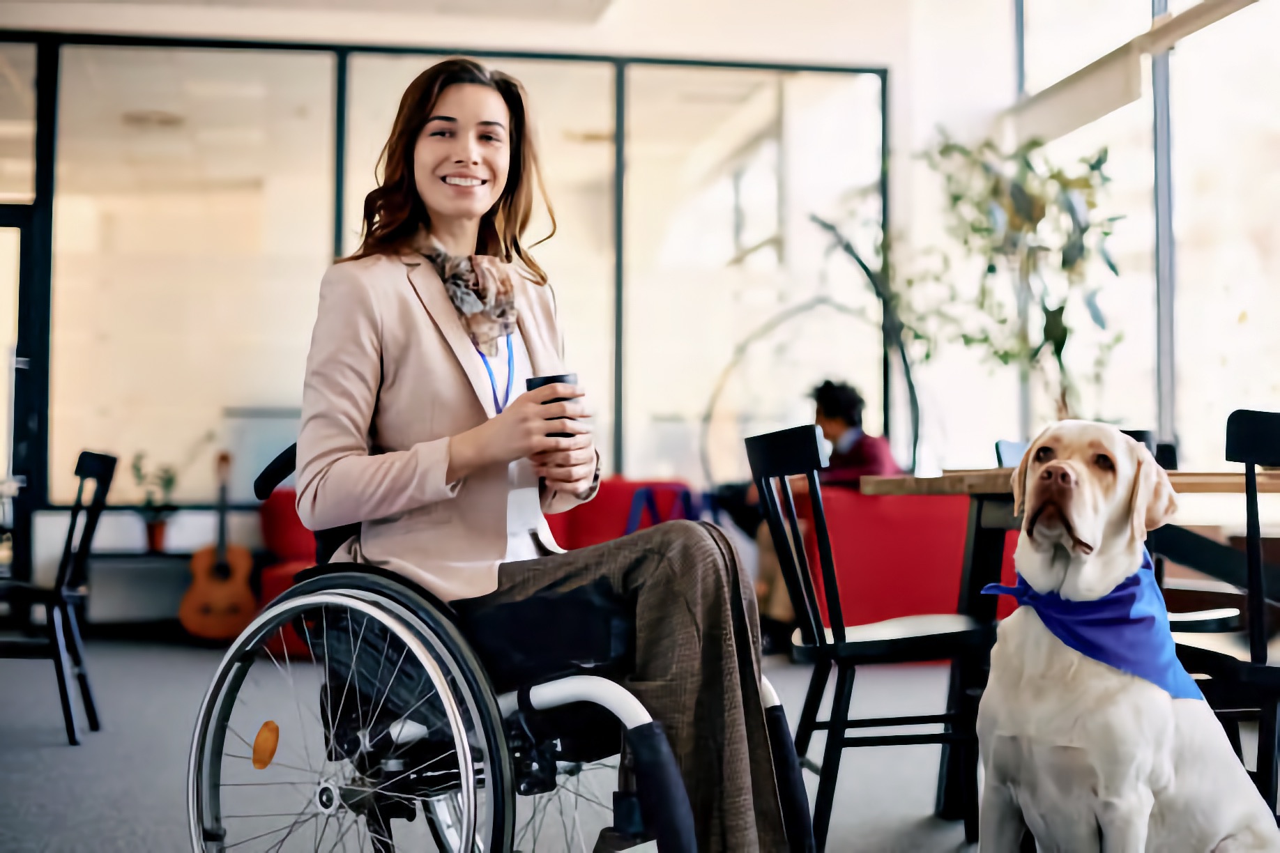 A young adult woman who uses a manual wheelchair sits indoors holding a drink and smiling toward the camera. Beside her sits a yellow Labrador service dog wearing a blue bandana, facing forward and alert.