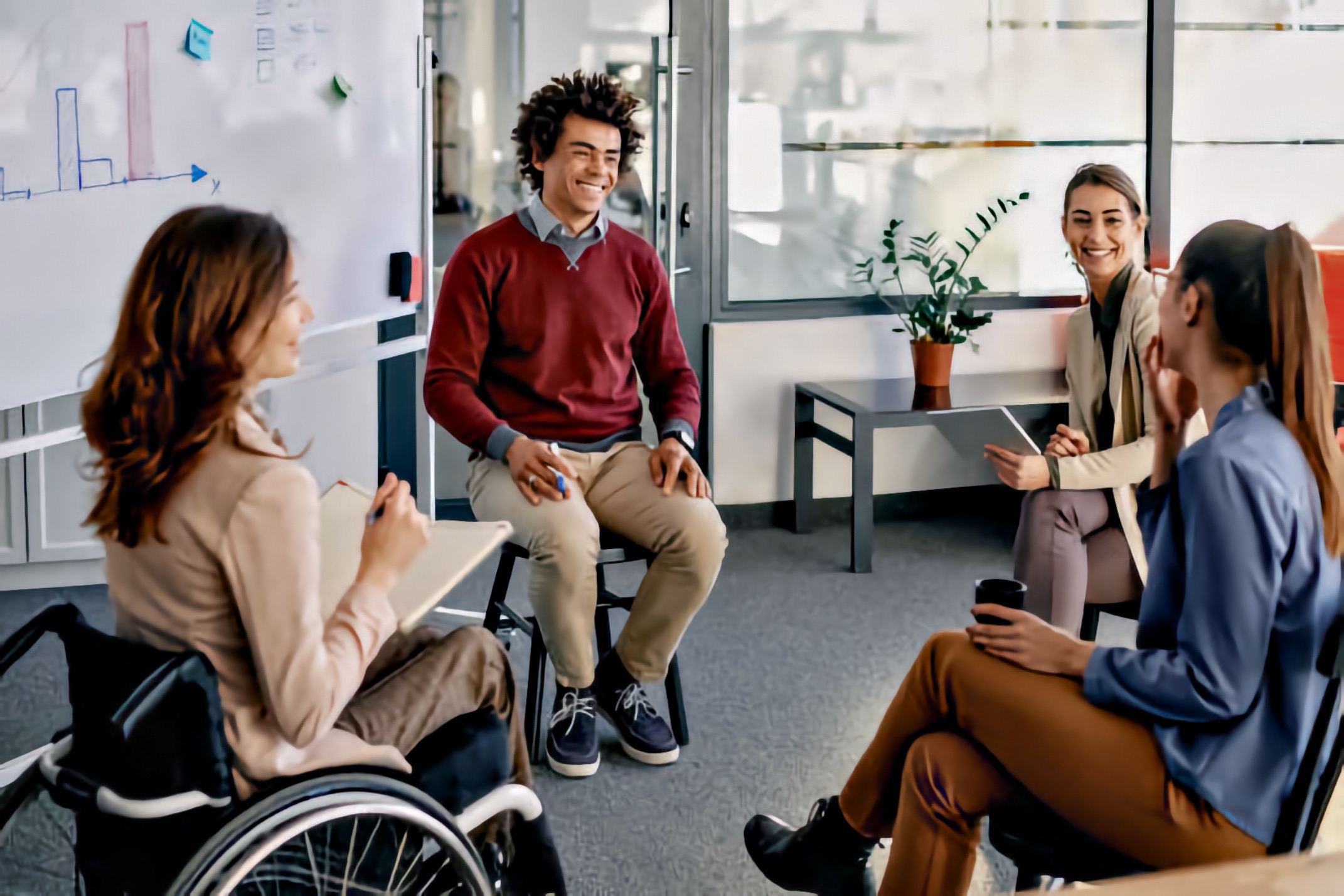 Four young adults sit in a circle in a meeting room having a group discussion. A young adult white woman who uses a manual wheelchair takes notes while facing the group. A young adult Black man sits smiling at the center, while two young adult white women sit opposite him listening and holding a tablet and a mug. A whiteboard and glass walls frame the accessible meeting space.